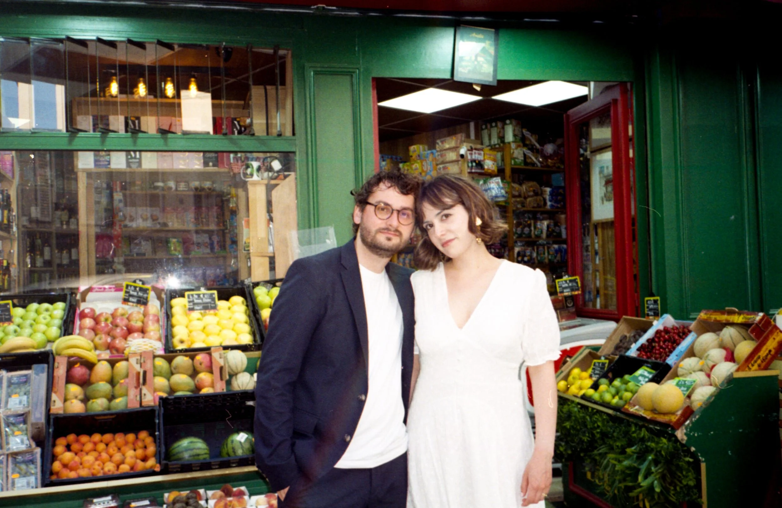 Deux mariés. Un homme avec des lunettes et une barbe, et une femme avec des cheveux bruns, posent devant un étal de fruits dans un marché ou une magasin, avec des pommes, melons et autres fruits.
