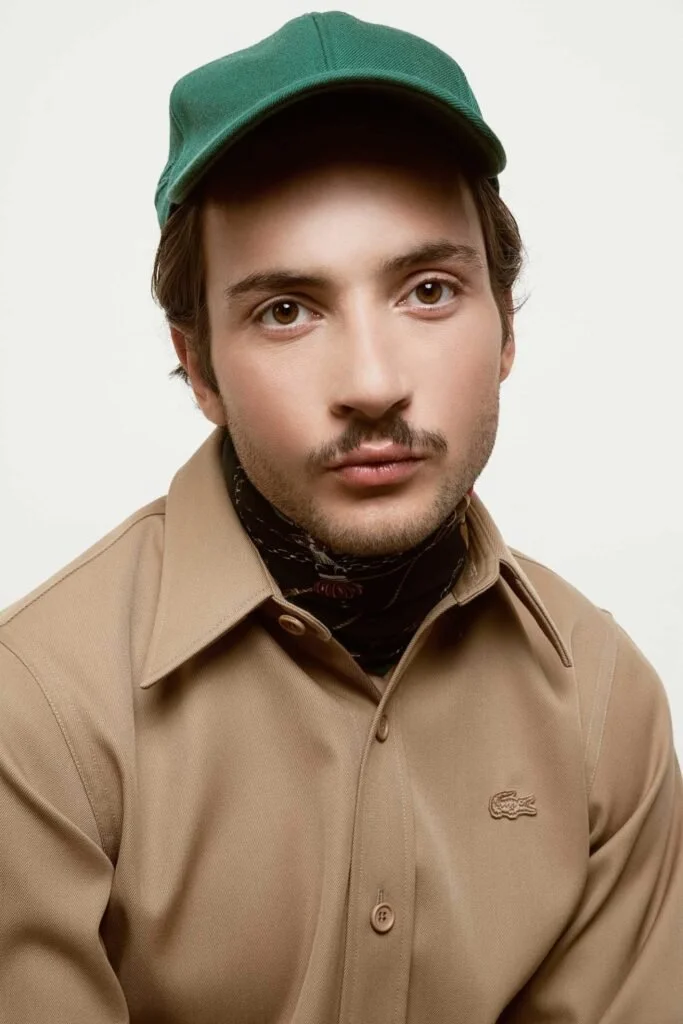 Un homme avec un chapeau vert, portant une chemise beige et un foulard noir autour du cou, regardant droit avec expression neutre.