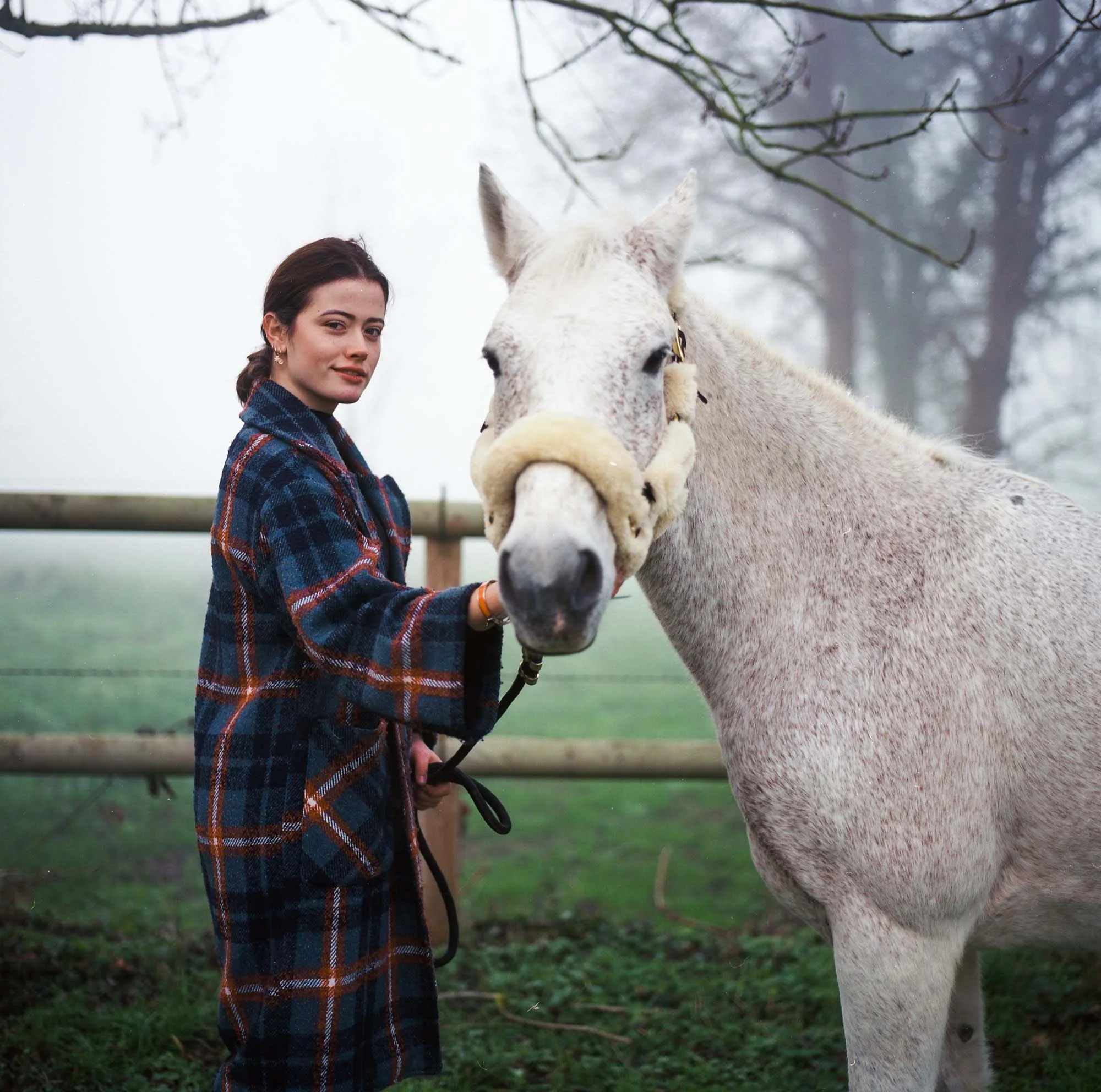 Jeune femme avec une écharpe à carreaux posant près d'un cheval blanc dans un champ verdoyant avec des arbres en arrière-plan lors d'une journée brumeuse.