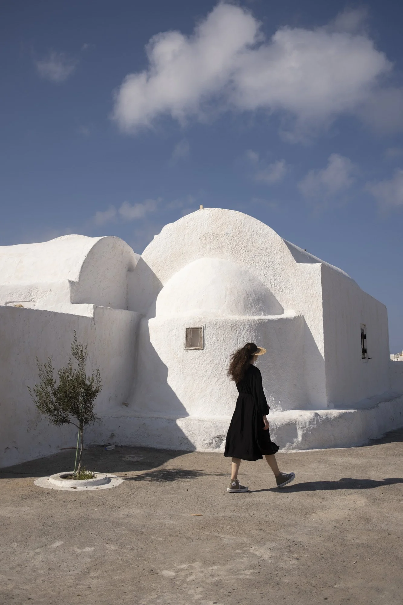 Une femme portant une robe noire et des baskets marche devant un bâtiment blanc aux formes arrondies, avec un ciel bleu et quelques nuages en arrière-plan à santorin