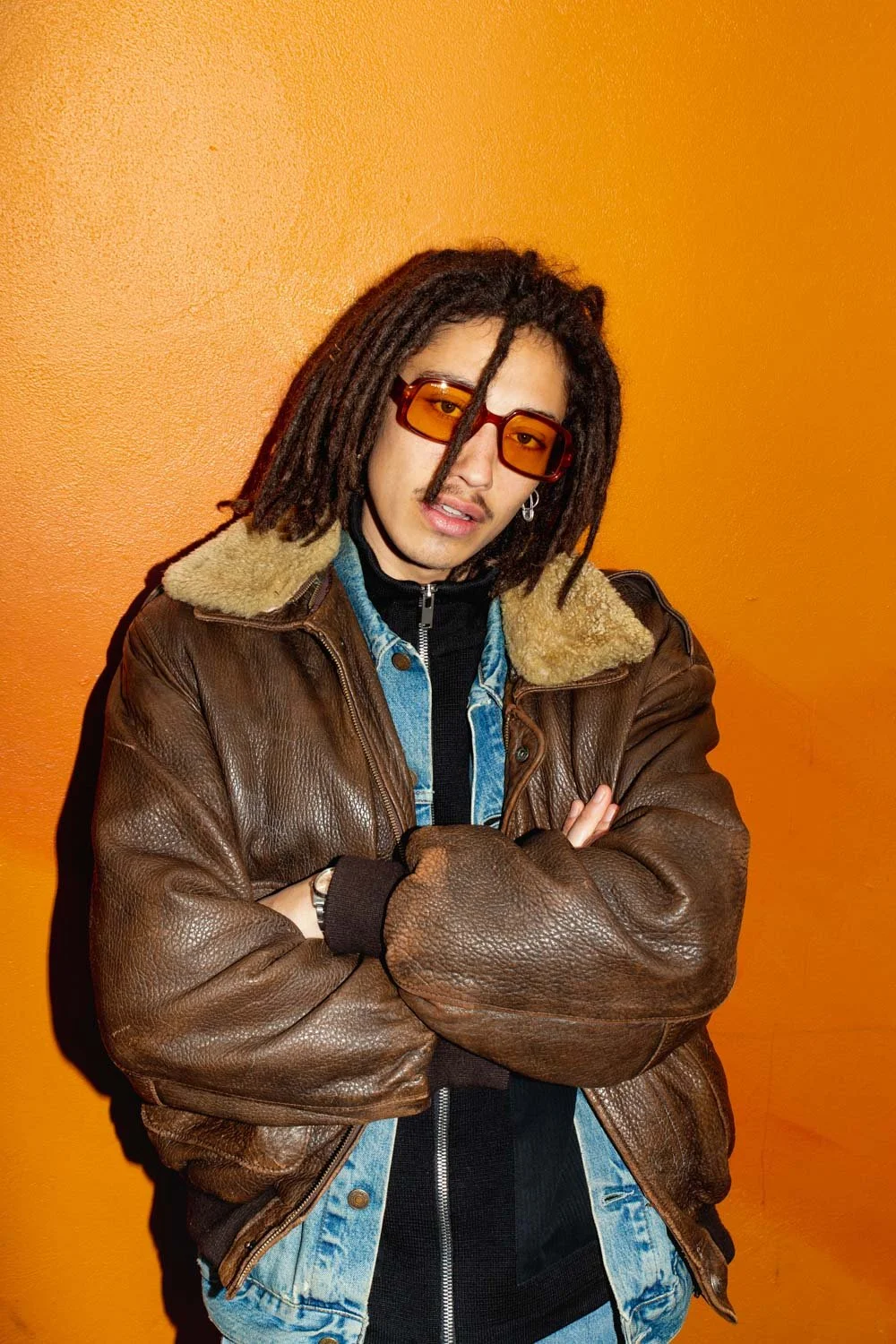 Un homme avec des dreadlocks, portant des lunettes orange, un blouson en cuir marron avec doublure en shearling, une veste en jean bleu et un haut noir, pose avec les bras croisés devant un mur orange.