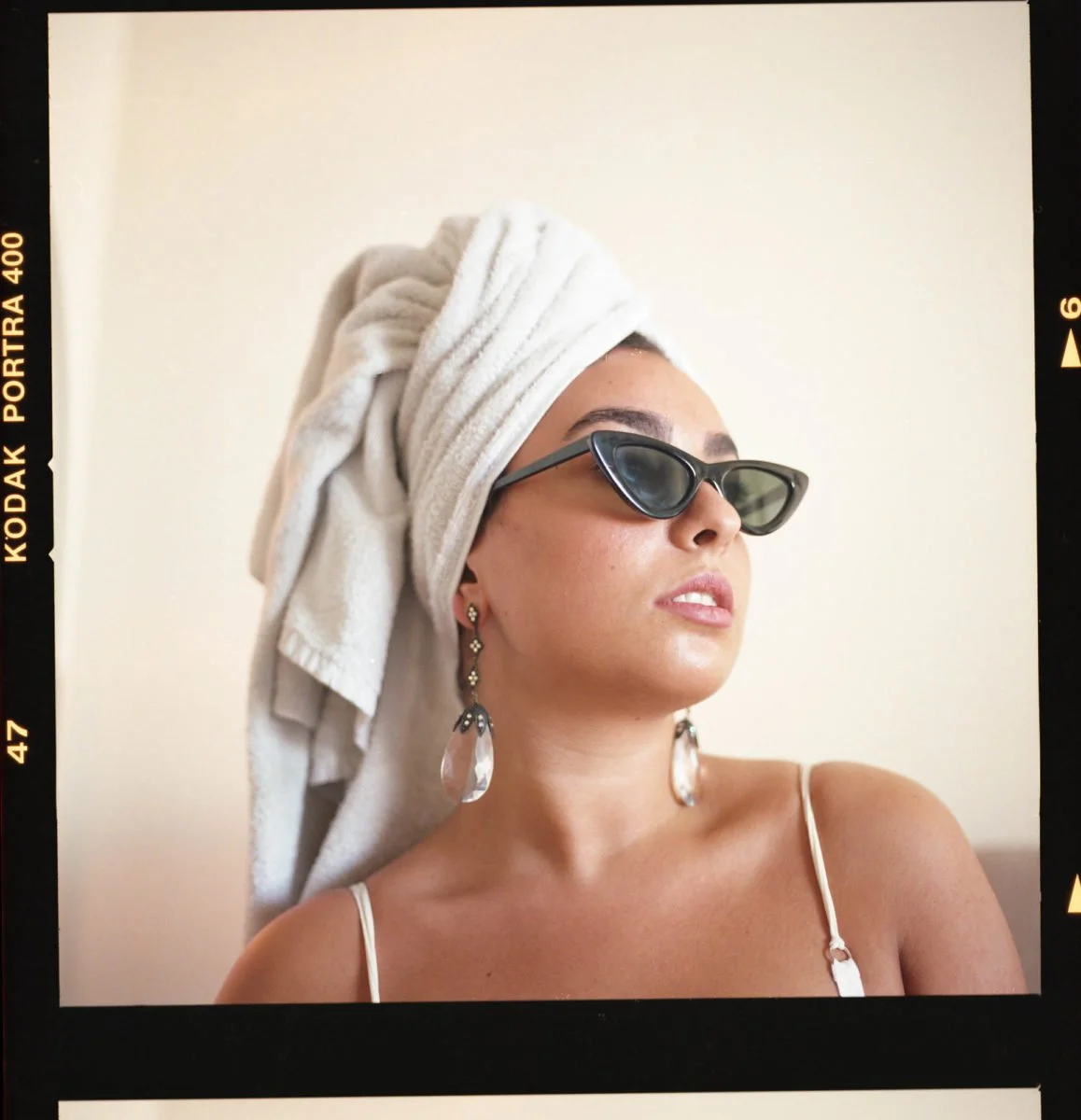 Une femme portant un turban blanc, des lunettes de soleil noires, des boucles d'oreilles pendantes et une robe à baillettes, vue de profil contre un fond clair.