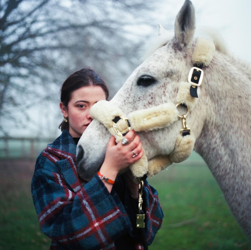 Une jeune femme tient la tête d'un cheval blanc avec des vêtements de peluche sur le nez et les oreilles dans un champ avec des arbres en arrière-plan.