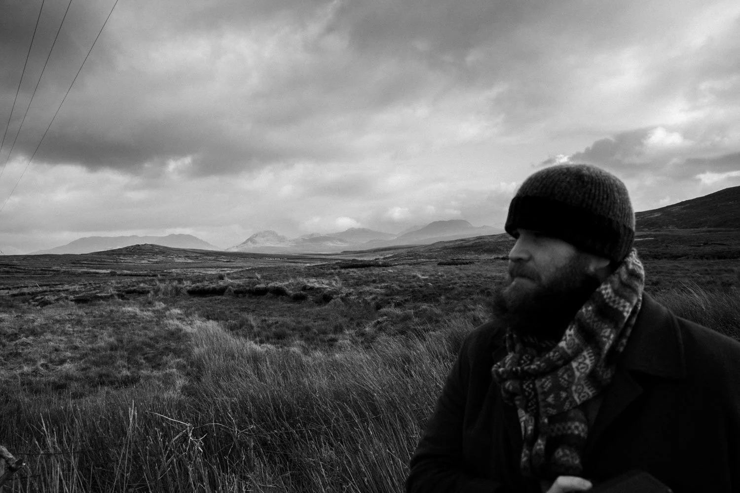 Un homme portant un bonnet et une écharpe regarde vers la droite dans un paysage de montagnes et de plaines sous un ciel nuageux, en noir et blanc.