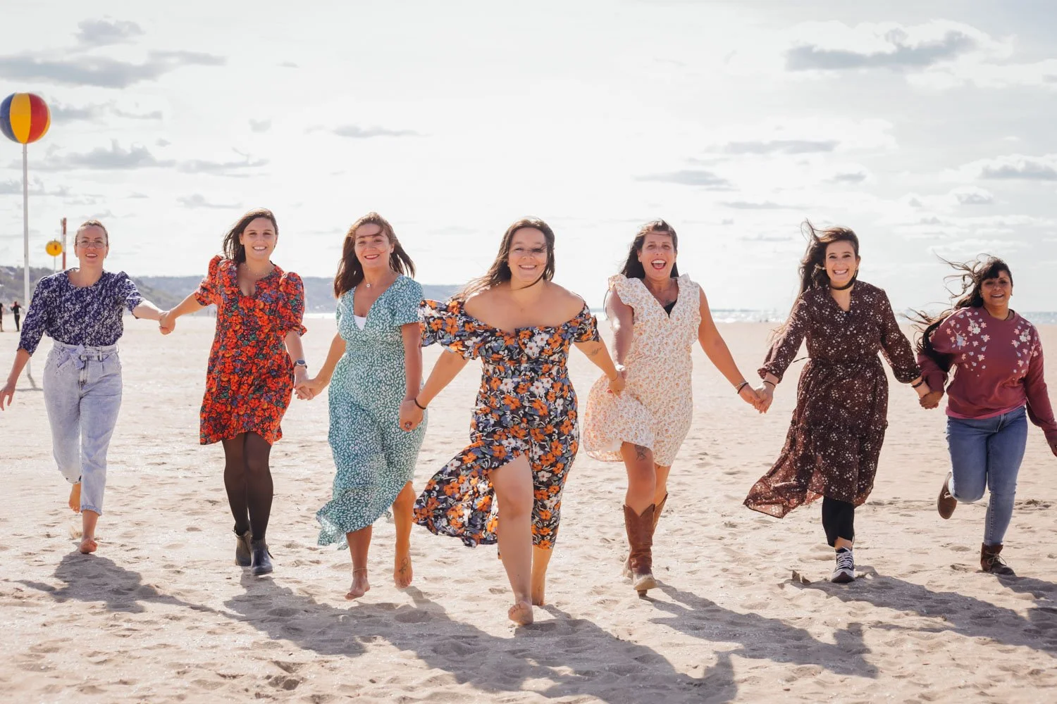 Groupe de huit femmes souriantes tenant la main et courant sur la plage, portant des robes d'été colorées.