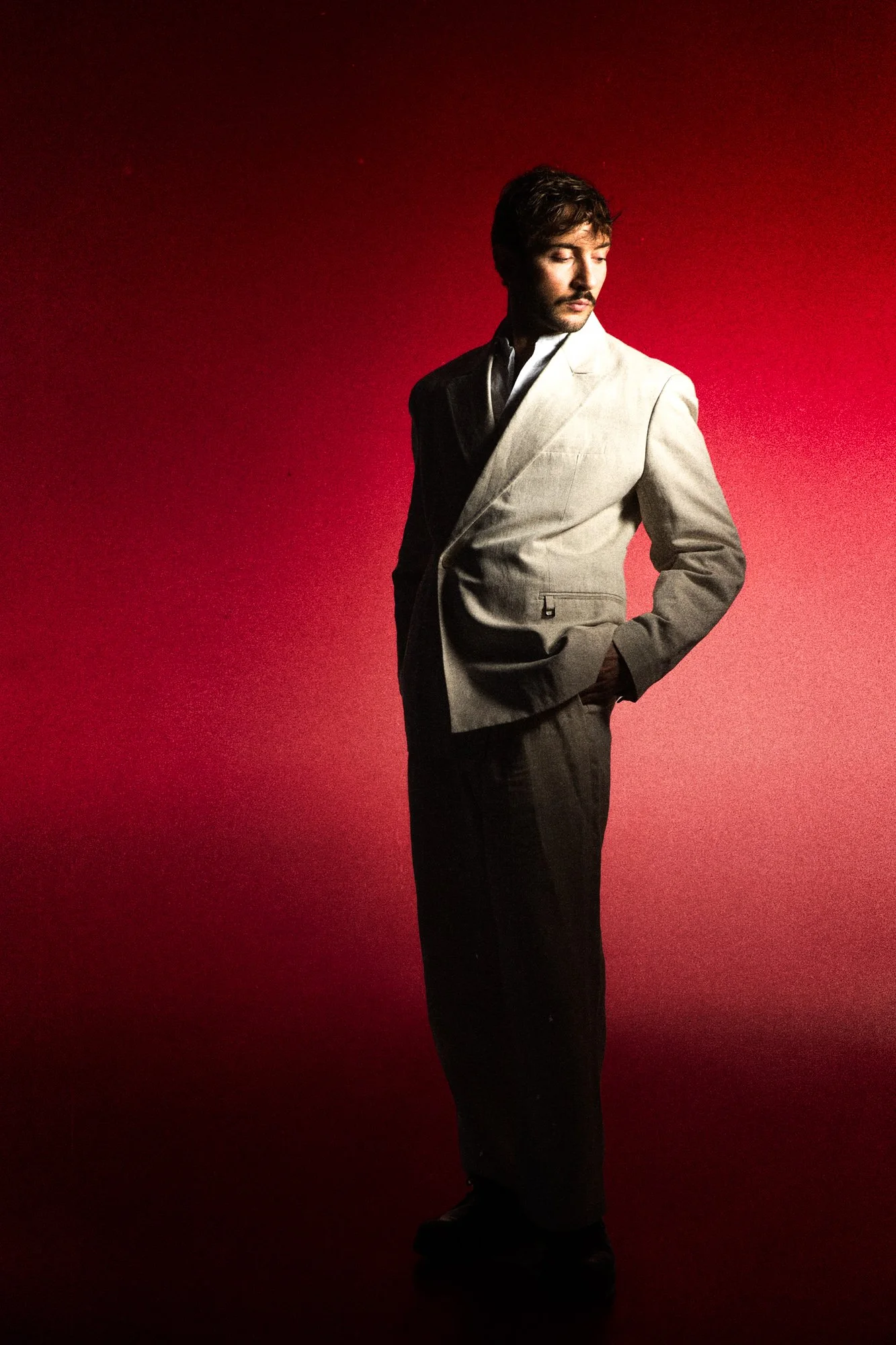 Homme en costume gris clair avec une veste et pantalon noir, posant avec regard vers la gauche, devant un fond rouge