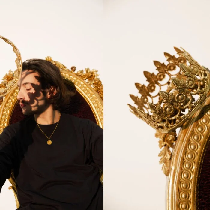 Un jeune homme avec des cheveux longs assis sur un trône doré, portant un collier or, avec une couronne dorée posée à côté.
