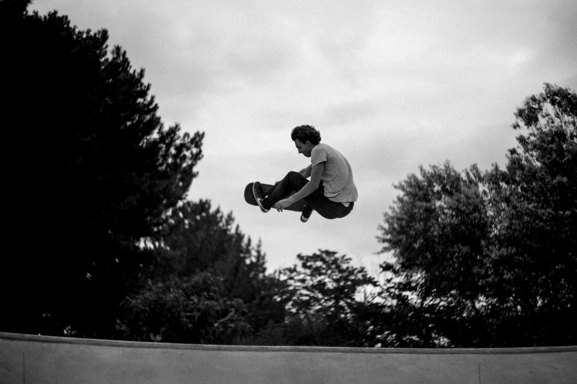 Un jeune homme en train de faire un saut avec sa planche de skate dans un parc, arbres en arrière-plan, ciel nuageux, en noir et blanc.