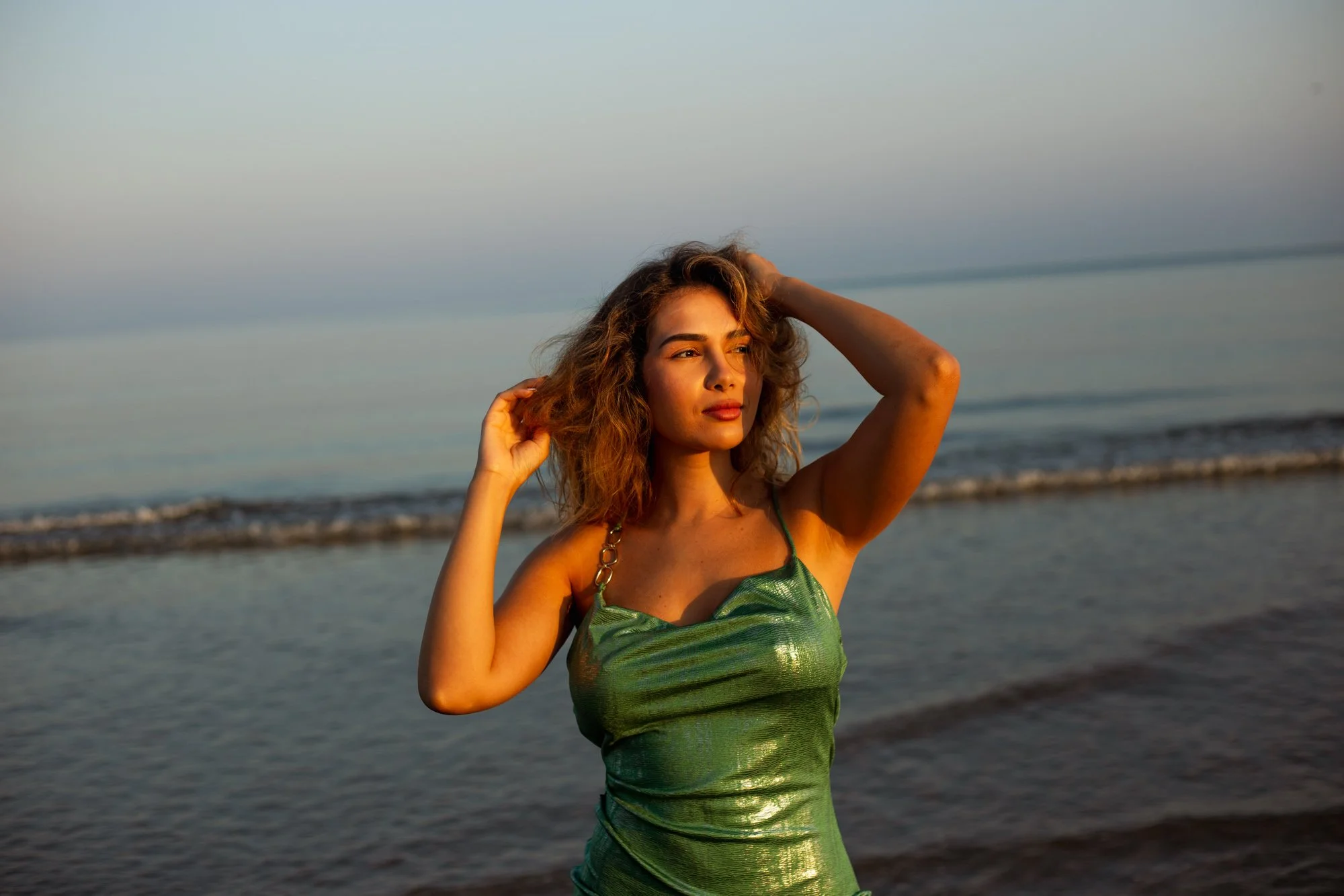 Une femme aux cheveux bouclés, portant une robe verte brillante, se tient à la plage avec la mer en arrière-plan, sous un ciel clair.