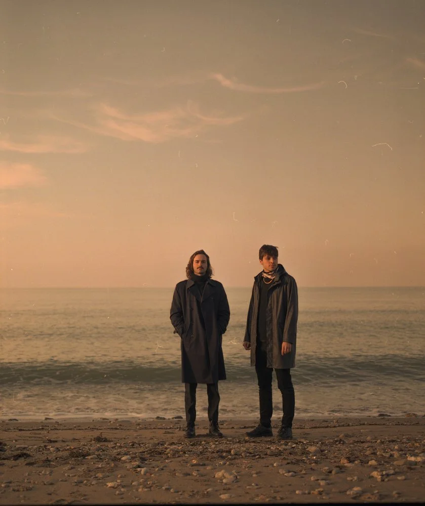 Deux jeunes hommes debout sur une plage au coucher du soleil, vêtus de manteaux longs, face à la mer calme.