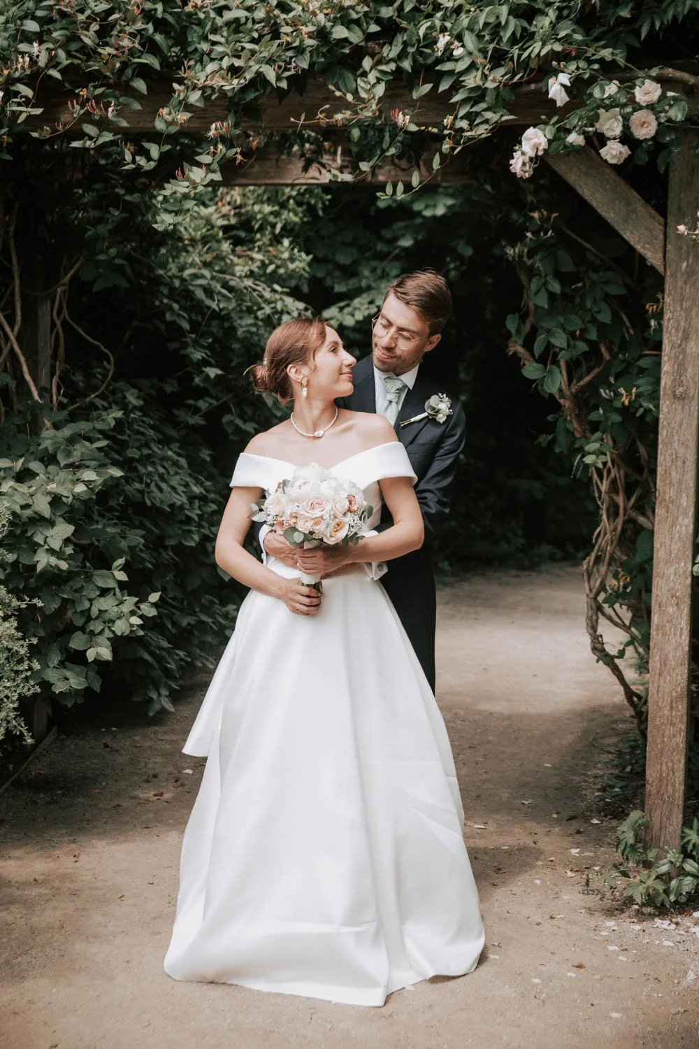 Un couple de mariés sous une arche de fleurs et de feuillage, le marié embrassant la mariée, dans un jardin.