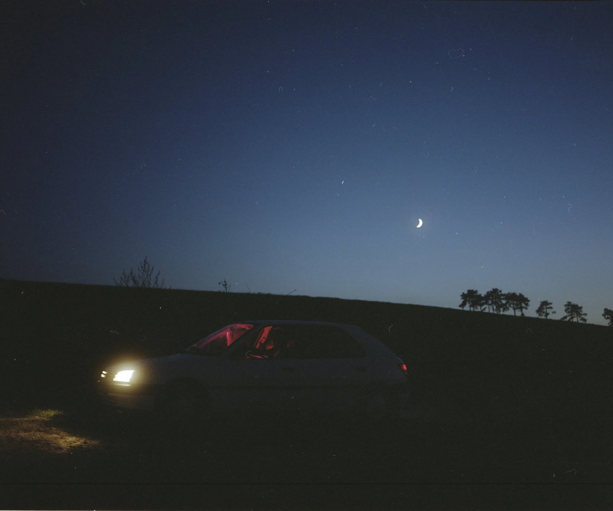 Voiture avec phares allumés dans un paysage nocturne, sous un ciel clair avec une lune en croissant et des étoiles. Photographie en argentique