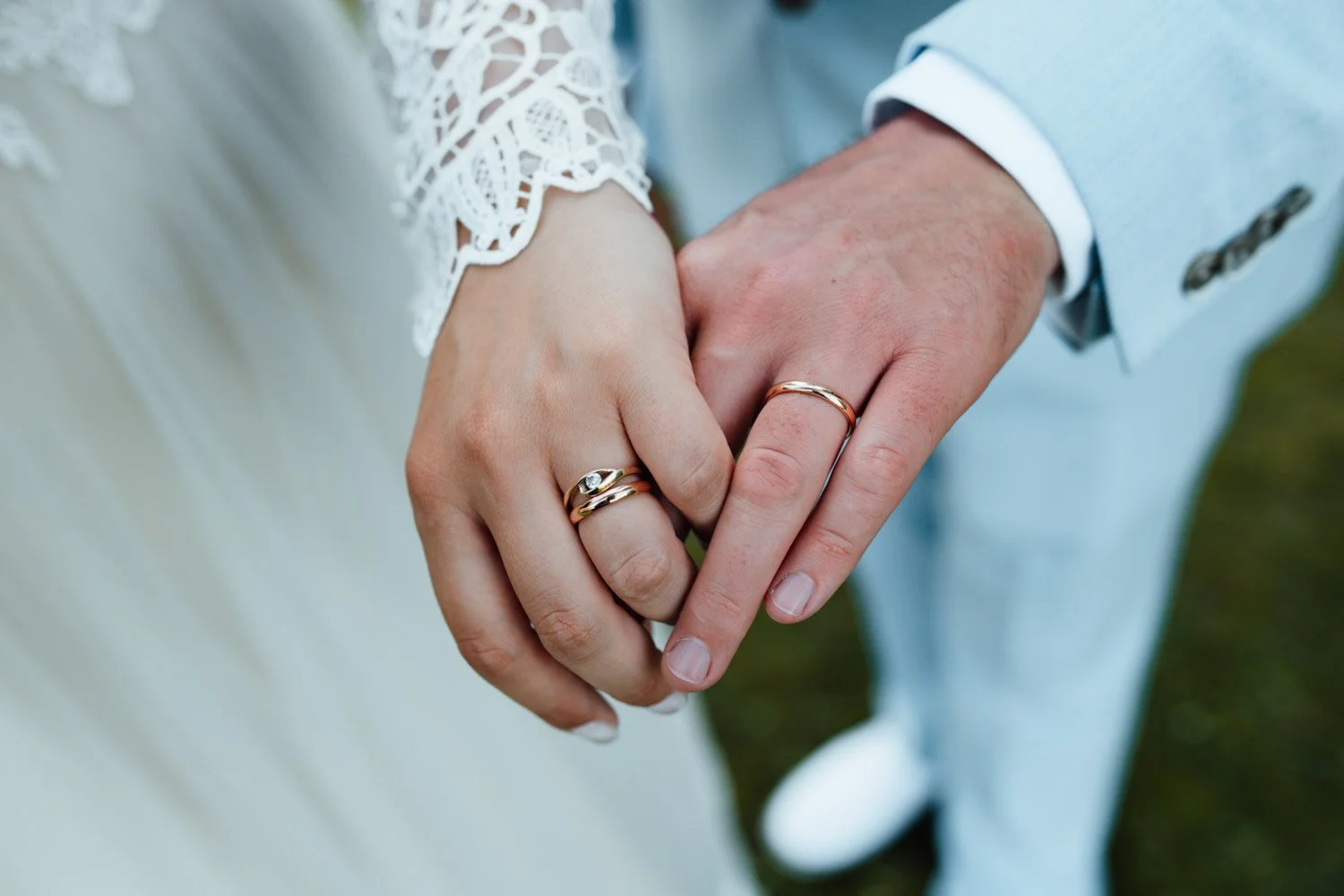 Mains d'un couple marié portant des alliances, la femme porte une robe avec des manches en dentelle, l'homme porte une veste bleu clair. La photo est prise en gros plan, montrant uniquement les mains et une partie de leur tenue.