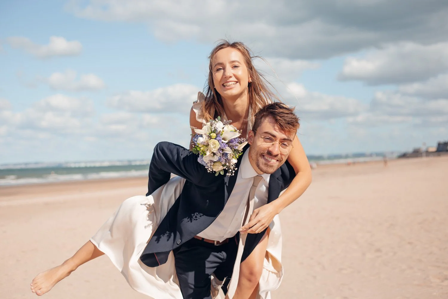 Un couple de mariage à la plage, la femme est sur le dos de l'homme, tous deux souriants, avec le ciel bleu et des nuages en arrière-plan.