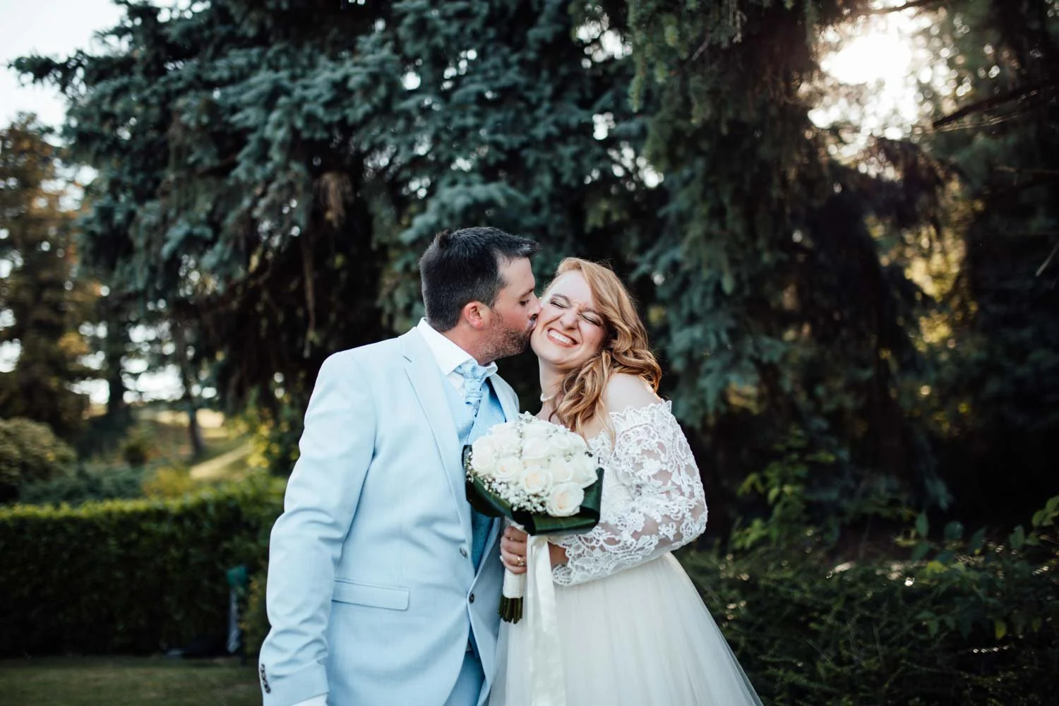 Un couple de mariage, l'homme embrassant la mariée sur la joue, la mariée souriant avec un bouquet de roses blanches, dans un parc avec un grand arbre en arrière-plan.