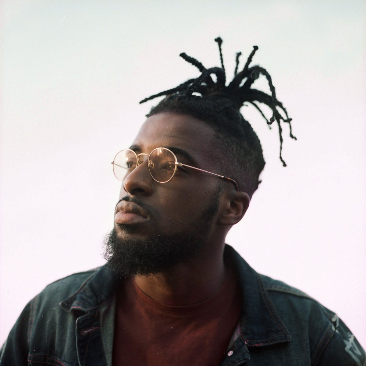 Portrait d'un homme avec des dreadlocks, portant des lunettes rondes, veste en denim, regardant vers la gauche.