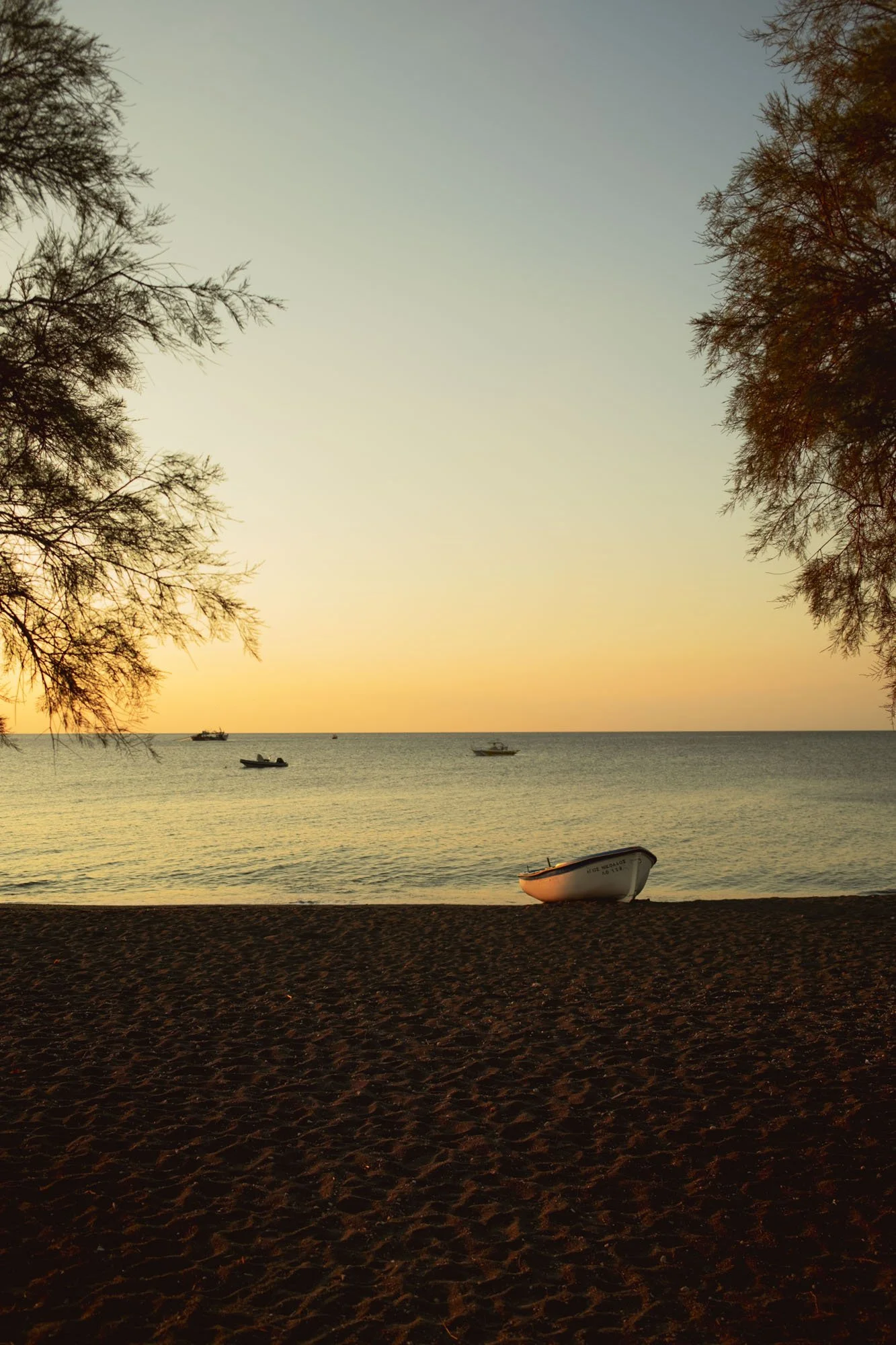 Coucher de soleil sur la mer avec des bateaux, arbres de part et d'autre, une barque échouée sur la plage à santorin
