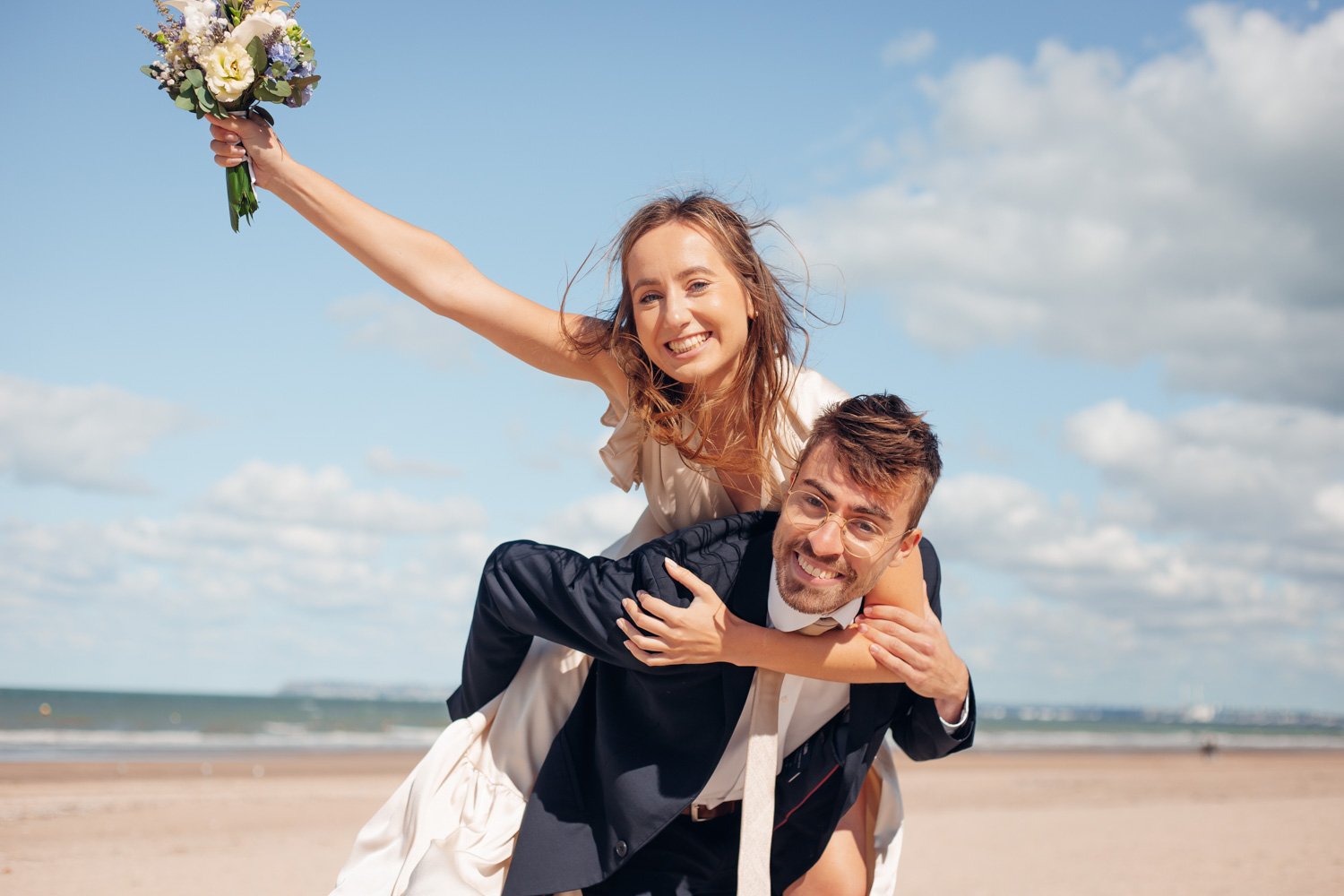 Un couple souriant sur la plage, la femme tenant un bouquet de fleurs et l'homme portant la femme sur ses épaules.