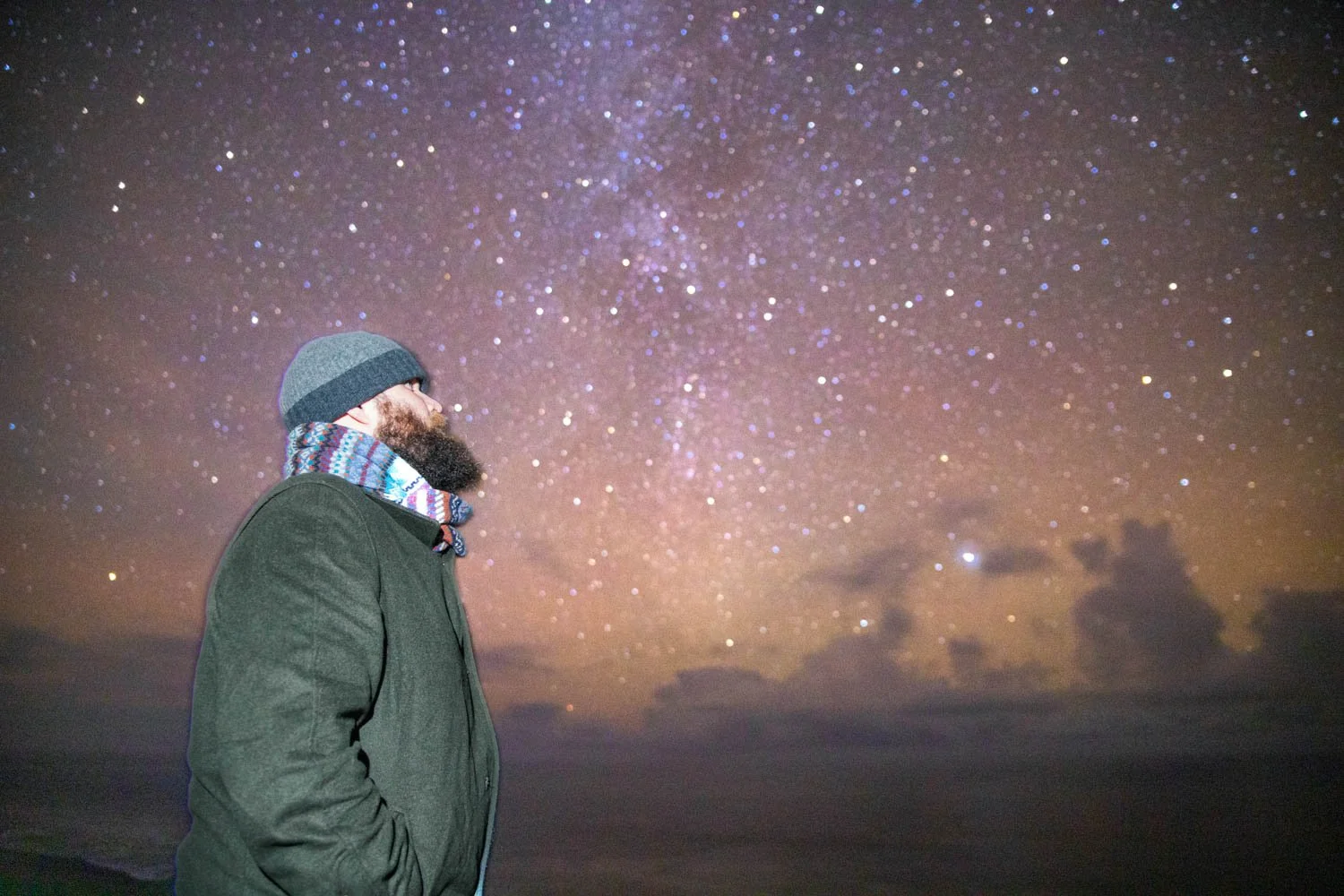 Un homme avec un manteau noir, un bonnet gris, et une écharpe colorée, regarde un ciel étoilé.