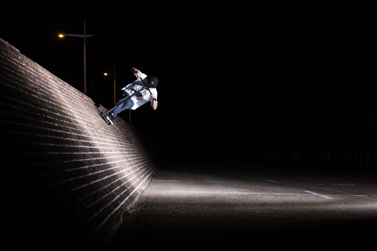 Jeune homme faisant du skate la nuit, éclairé par une lampe au sol, avec un mur en briques en pente et un ciel noir en arrière-plan.