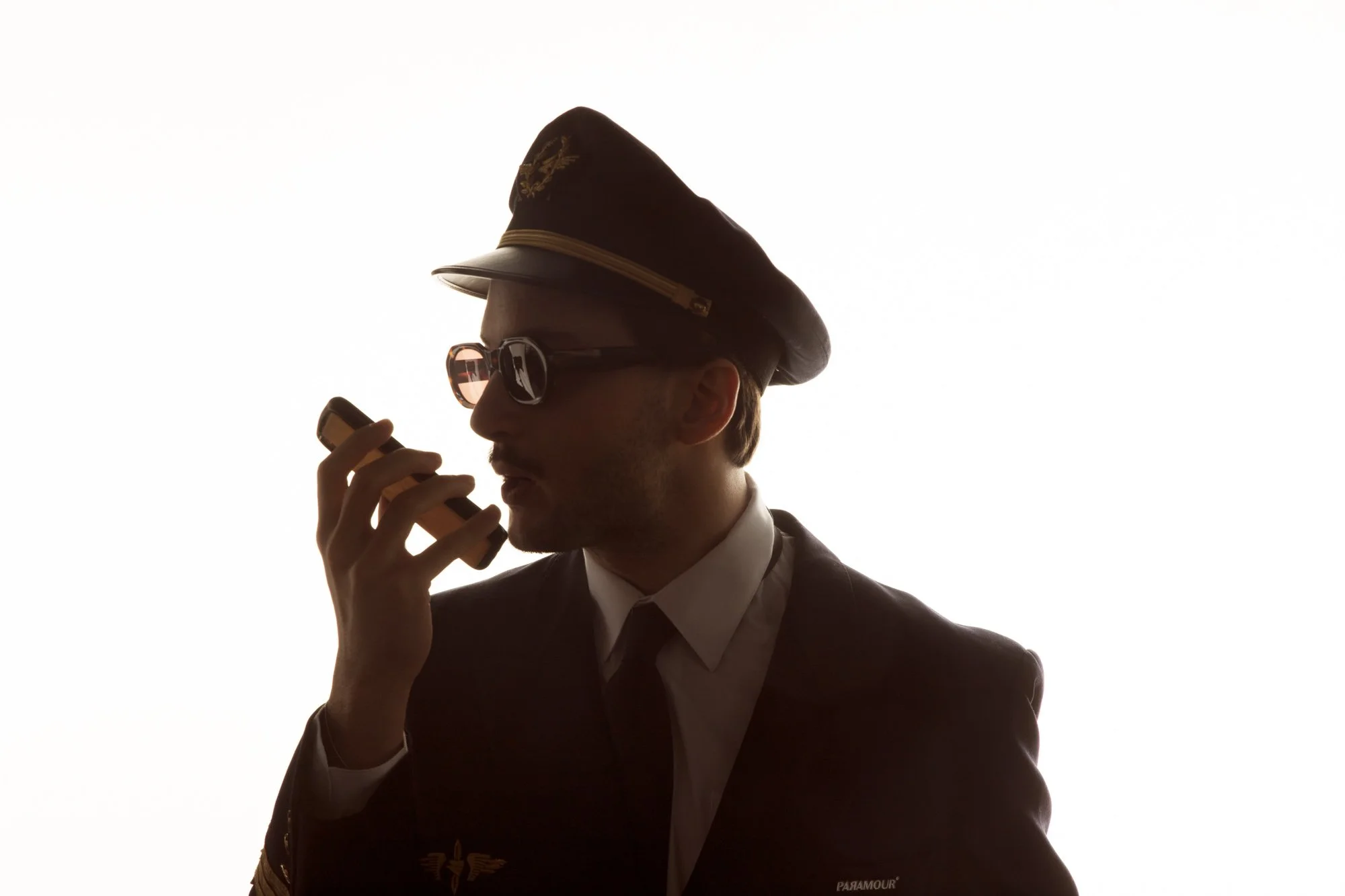 Lord Esperanza en costume noir et lunettes de soleil porte un chapeau de pilote et utilise un téléphone portable, silhouetté contre un arrière-plan lumineux.