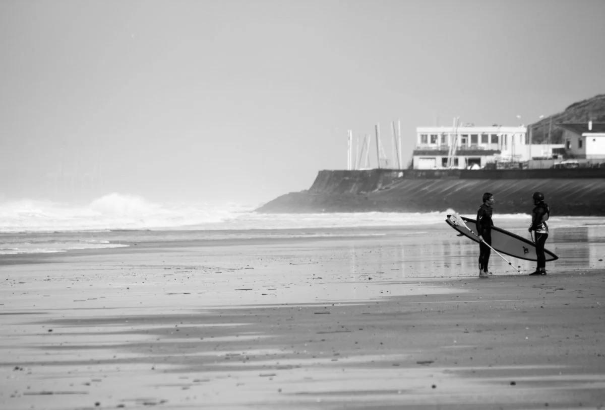 Deux personnes se tiennent avec leur planche de surf sur une plage, discutant, avec des bâtiments et des voiliers en arrière-plan.