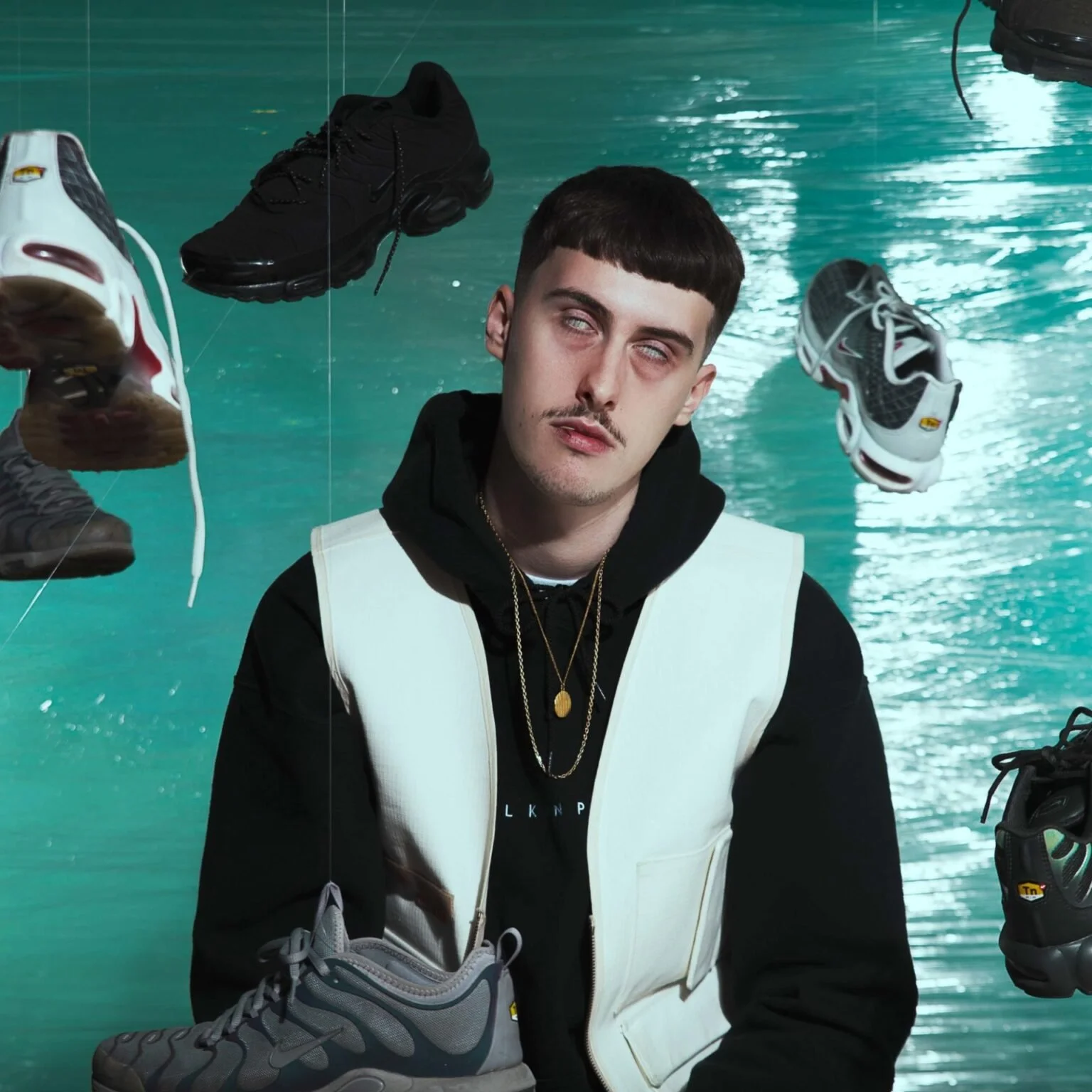 Un jeune homme avec une coupe au style moderne, portant un sweatshirt noir et une veste blanche, assis devant un mur vert, entouré de chaussures de sport suspendues.