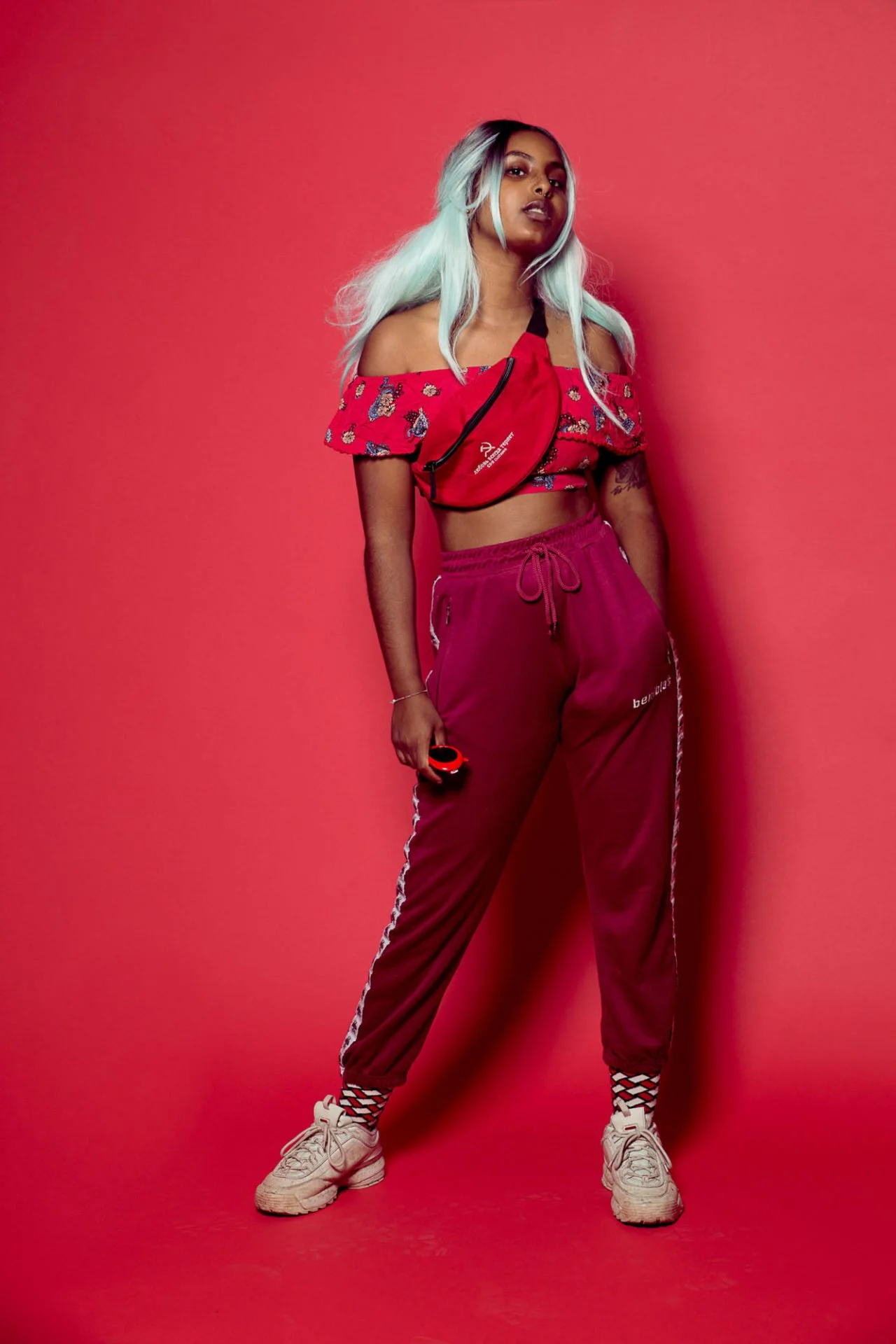 Jeune femme stylée avec cheveux longs décolorés, vêtu d'un crop top à épaules dénudées avec motif floral rouge, pantalon de survêtement violet avec rayures blanches sur le côté, chaussures blanches, et portant une banane rouge, se tenant contre un mu