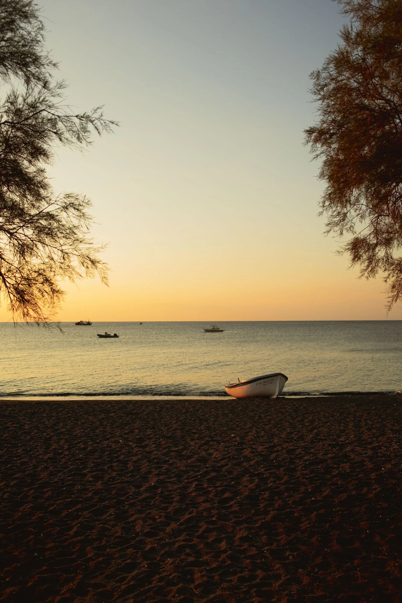 Coucher de soleil sur une plage avec un bateau abandonné sur le rivage, plusieurs bateaux en mer, arbres au bord de l'eau, ambiance calme et paisible à santorin