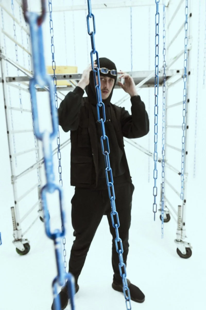 Homme portant un bonnet noir et des lunettes de natation, debout dans une structure en métal avec des chaînes bleues suspendues autour de lui.