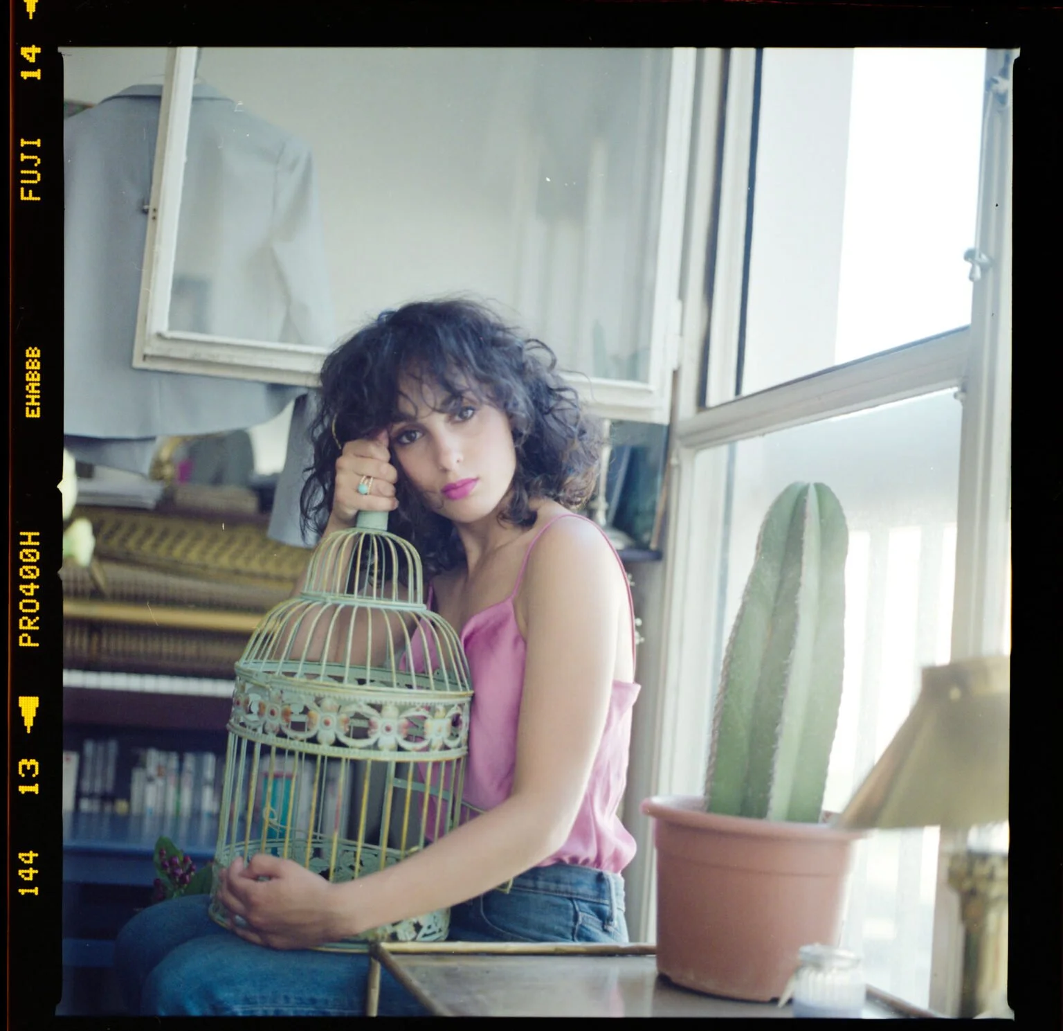 Une jeune femme aux cheveux bouclés et vêtue d'un top rose assise à une table, tenant une cage ornamentale, dans un intérieur bien éclairé avec un cactus dans un pot en terre cuite sur la table à côté d'elle.