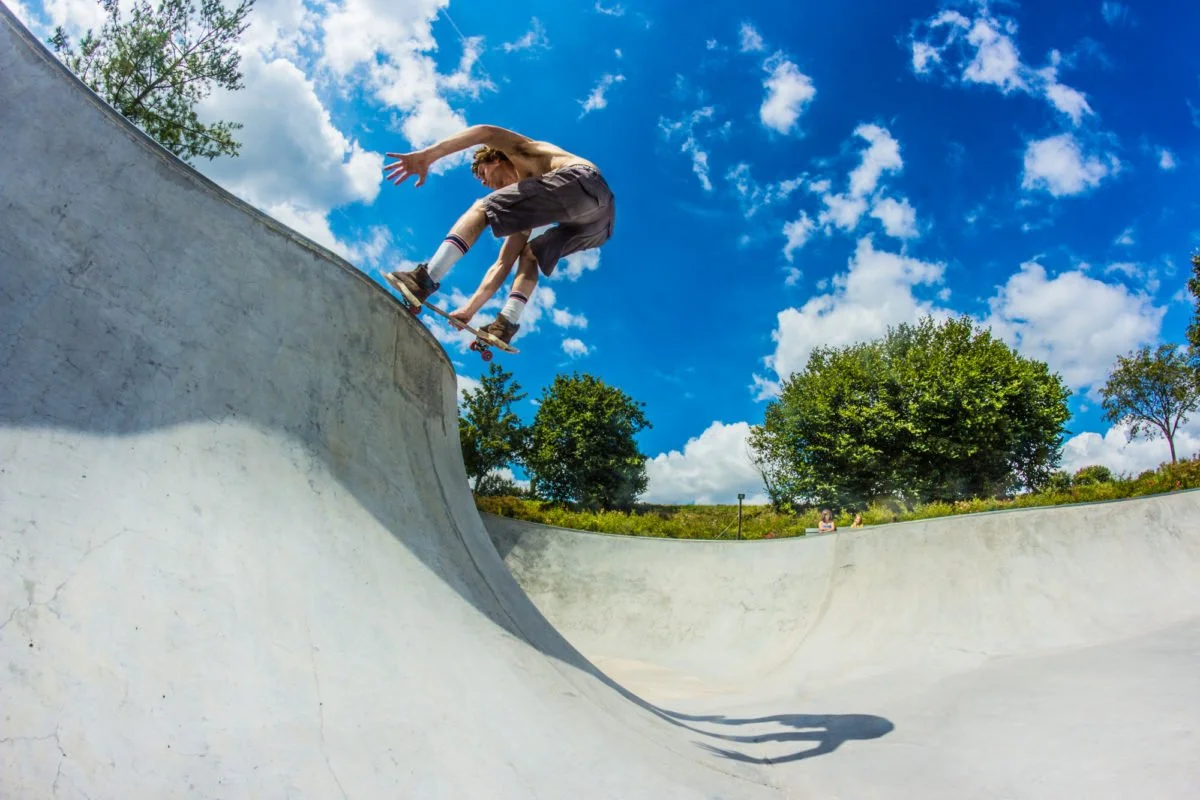 Jeune homme en skateboard effectuant un trick dans une rampe en béton sous un ciel bleu avec quelques nuages et des arbres en arrière-plan.