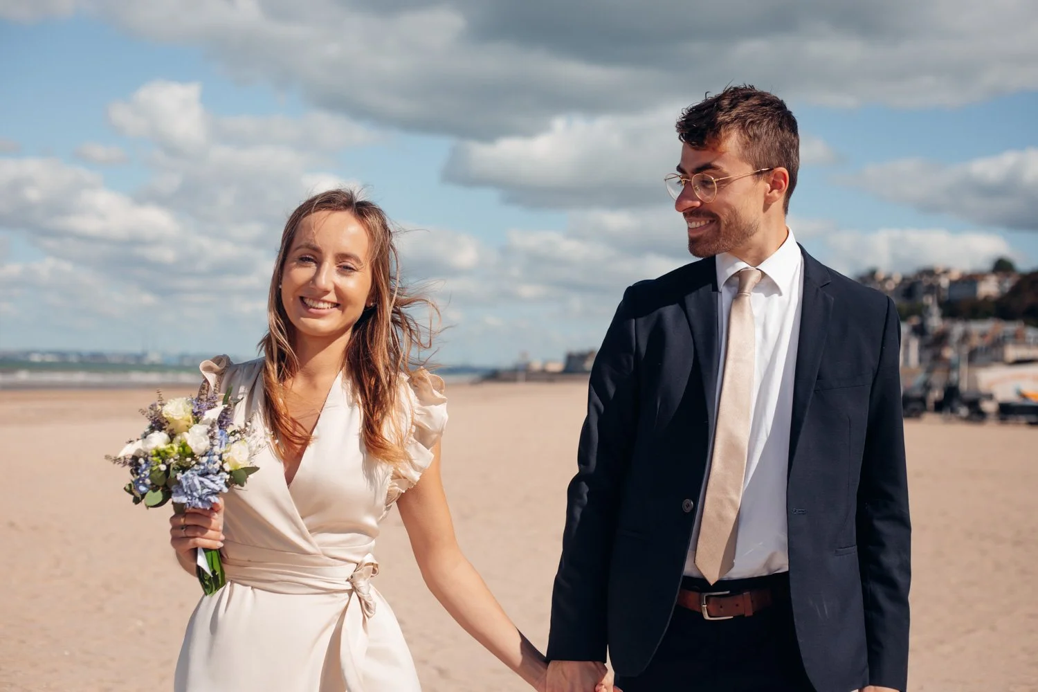 Un couple en mariage marche main dans la main sur la plage, la femme tient un bouquet de fleurs et porte une robe élégante, l'homme porte un costume sombre avec une cravate claire, le ciel est partiellement nuageux.