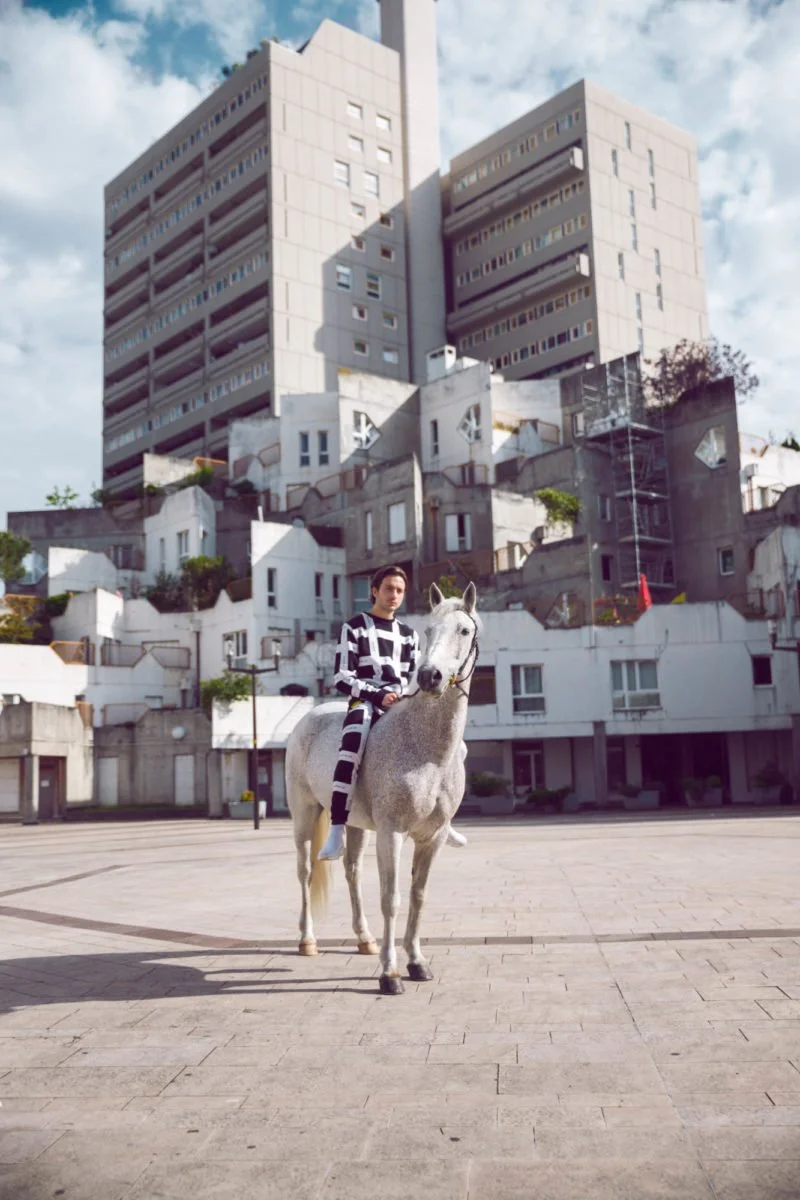 Une femme en tenue noire et blanche monte un cheval blanc dans une place urbaine, avec un grand immeuble moderne en arrière-plan et un ciel partiellement nuageux.