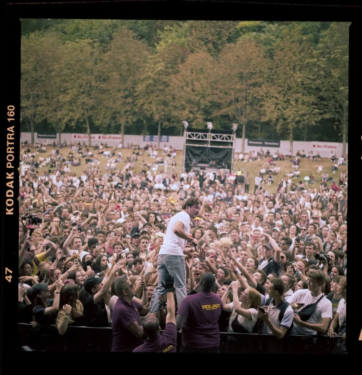 Une foule de personnes assistant à un concert en plein air, avec un chanteur au centre interagissant avec le public, entouré de sécurité.