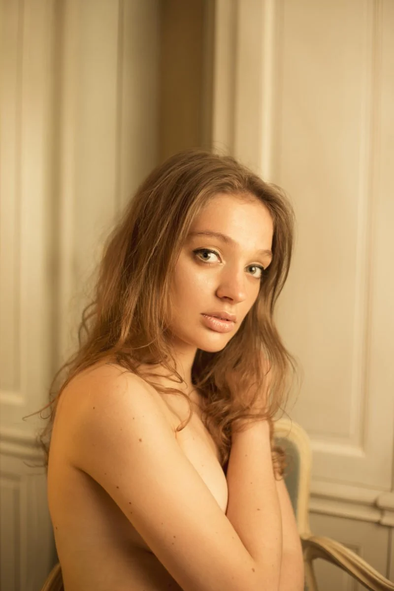 Jeune femme avec cheveux bruns et yeux bleus, posant nue dans une pièce avec des murs en bois clairs.