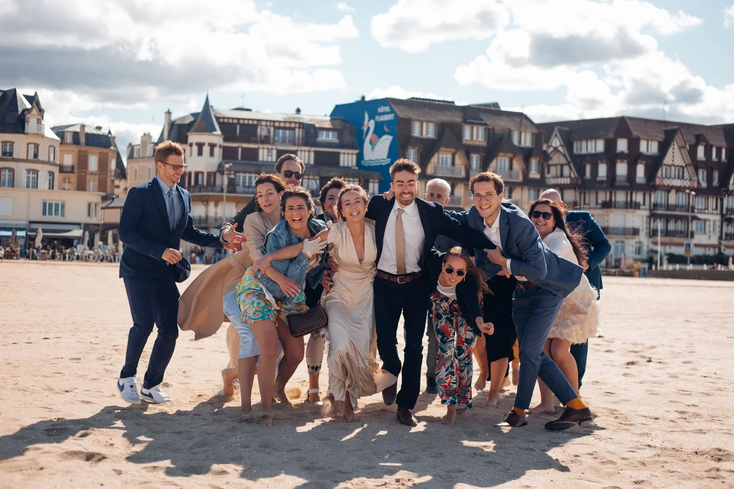 Groupe de personnes souriantes en tenues décontractées et formelles sur la plage, posant joyeusement pour une photo en famille ou entre amis, avec des bâtiments résidentiels en arrière-plan.