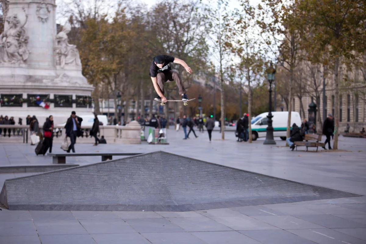 Joseph Garbaccio en plein saut au-dessus d'une structure urbaine à république, avec des arbres et des personnes qui se promènent en arrière-plan, dans une ville en automne.