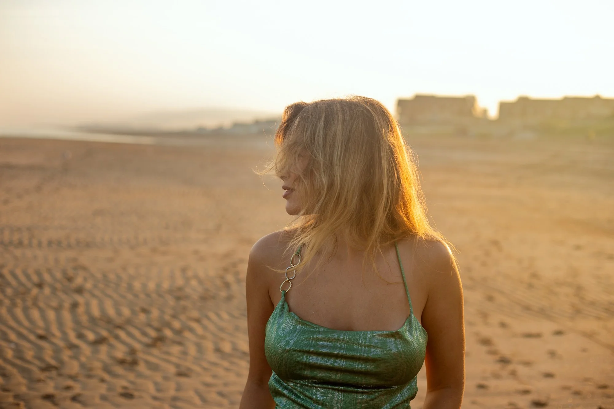 Une femme aux cheveux longs et blonds portant une robe verte à bretelles, debout sur une plage au coucher du soleil.