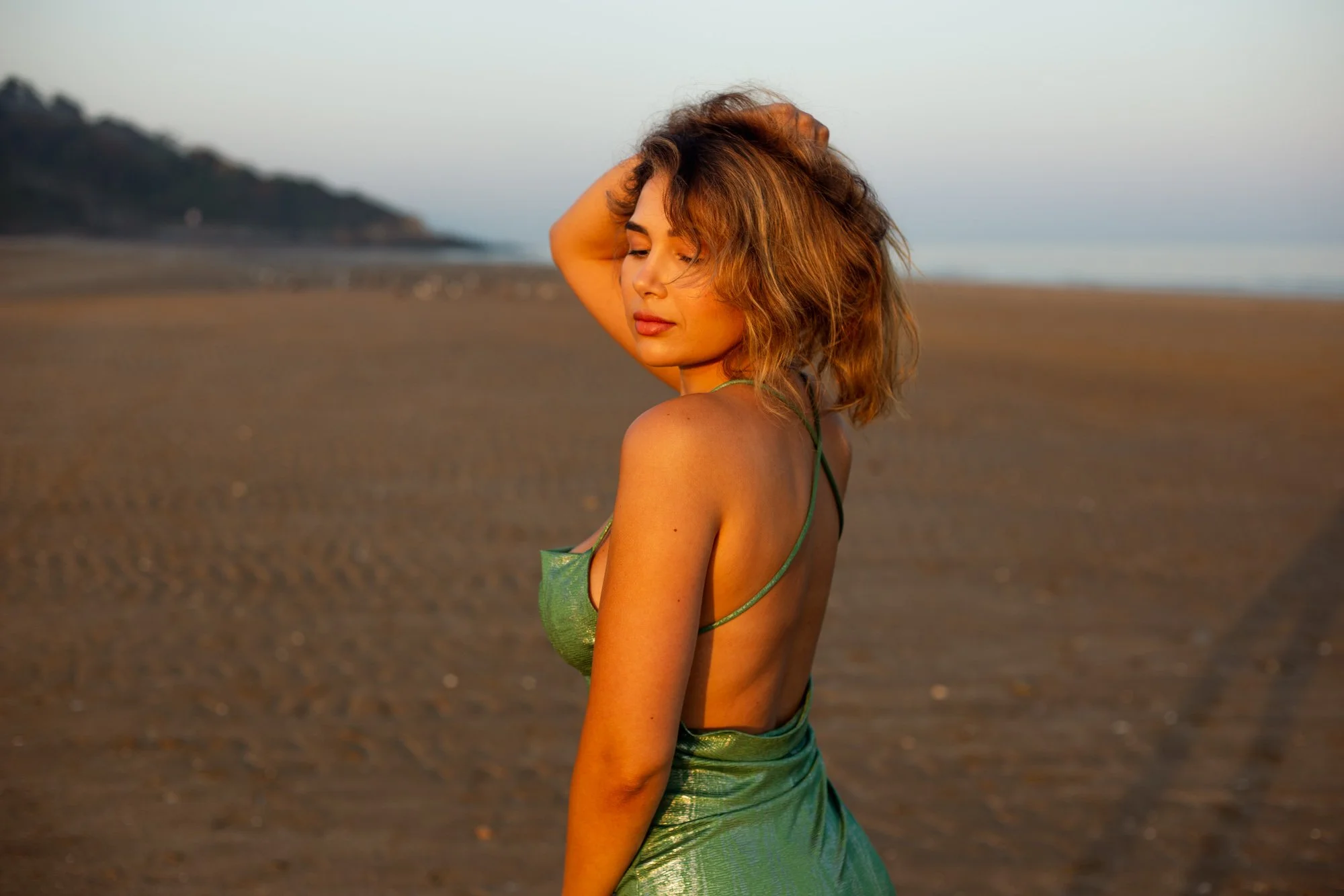 Une femme avec des cheveux bouclés et bruns pose sur la plage, portant une robe verte en satin, au coucher du soleil.