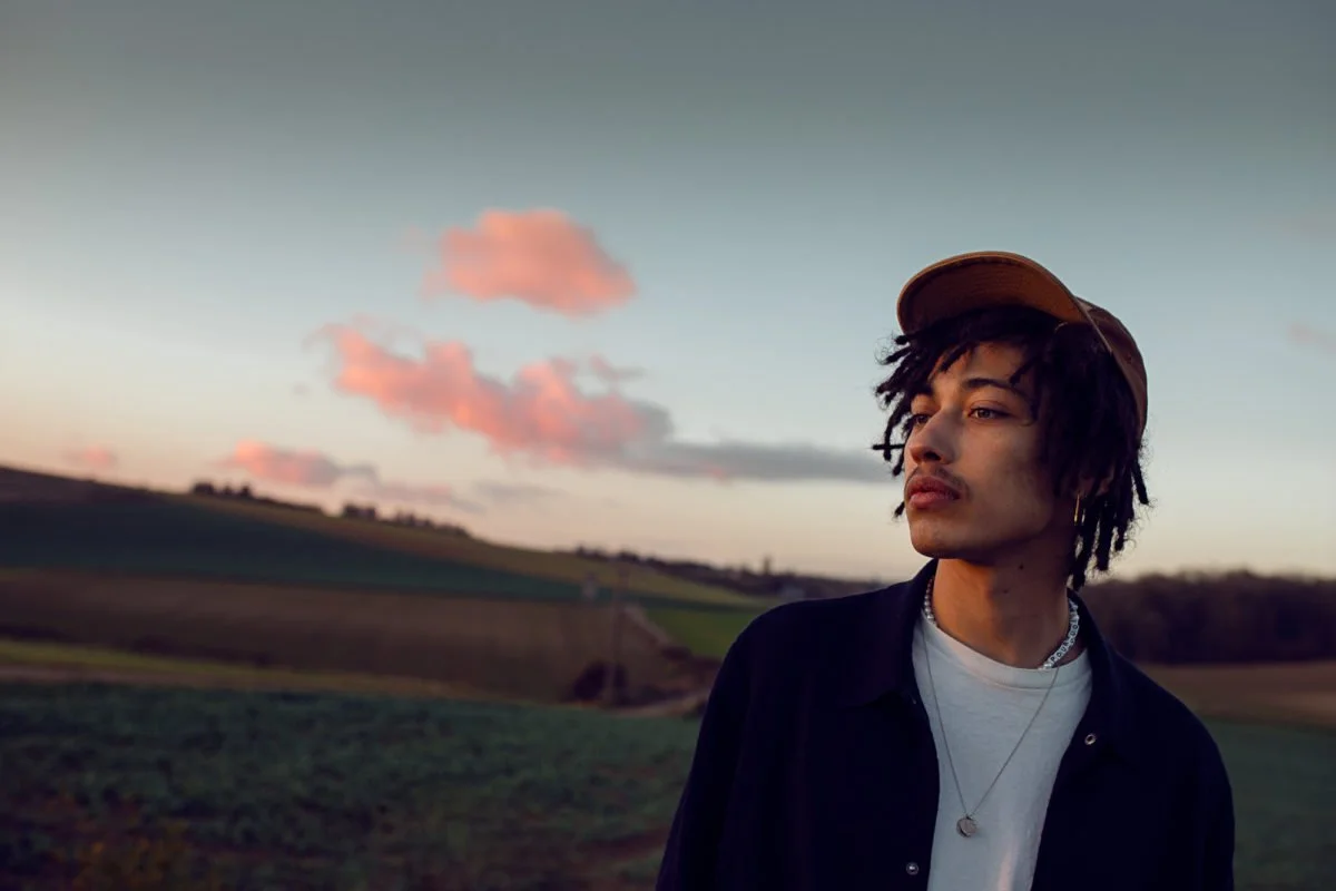 Un jeune homme avec des cheveux en dreadlocks portant une casquette, une chaîne et un collier, se tient dans un champ lors du coucher du soleil.