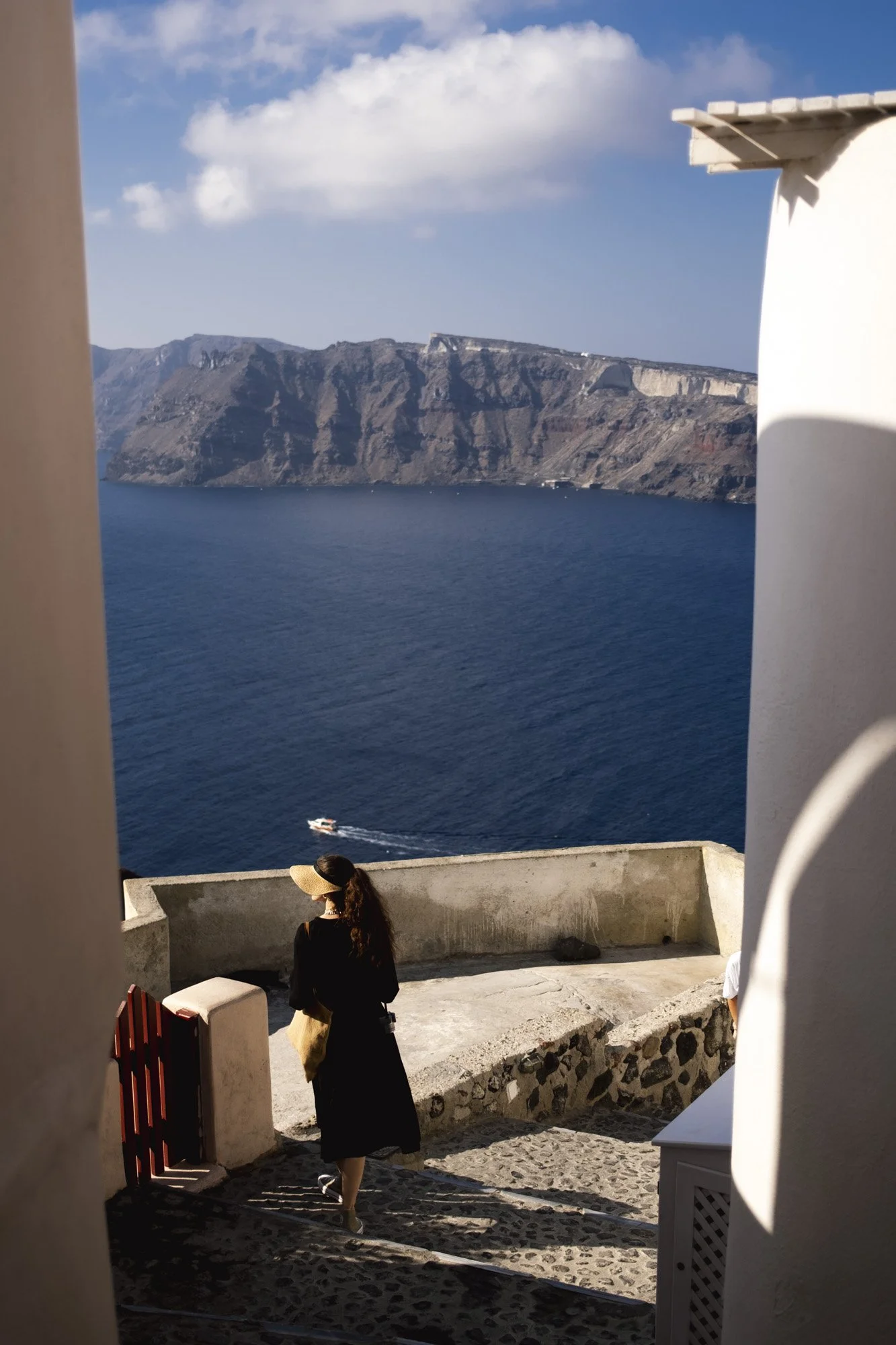 Une femme portant un chapeau en osier et une robe noire qui marche le long d'une terrasse à Santorin, avec des falaises jaunes et la mer bleue à l'arrière-plan, sous un ciel partiellement nuageux à santorin