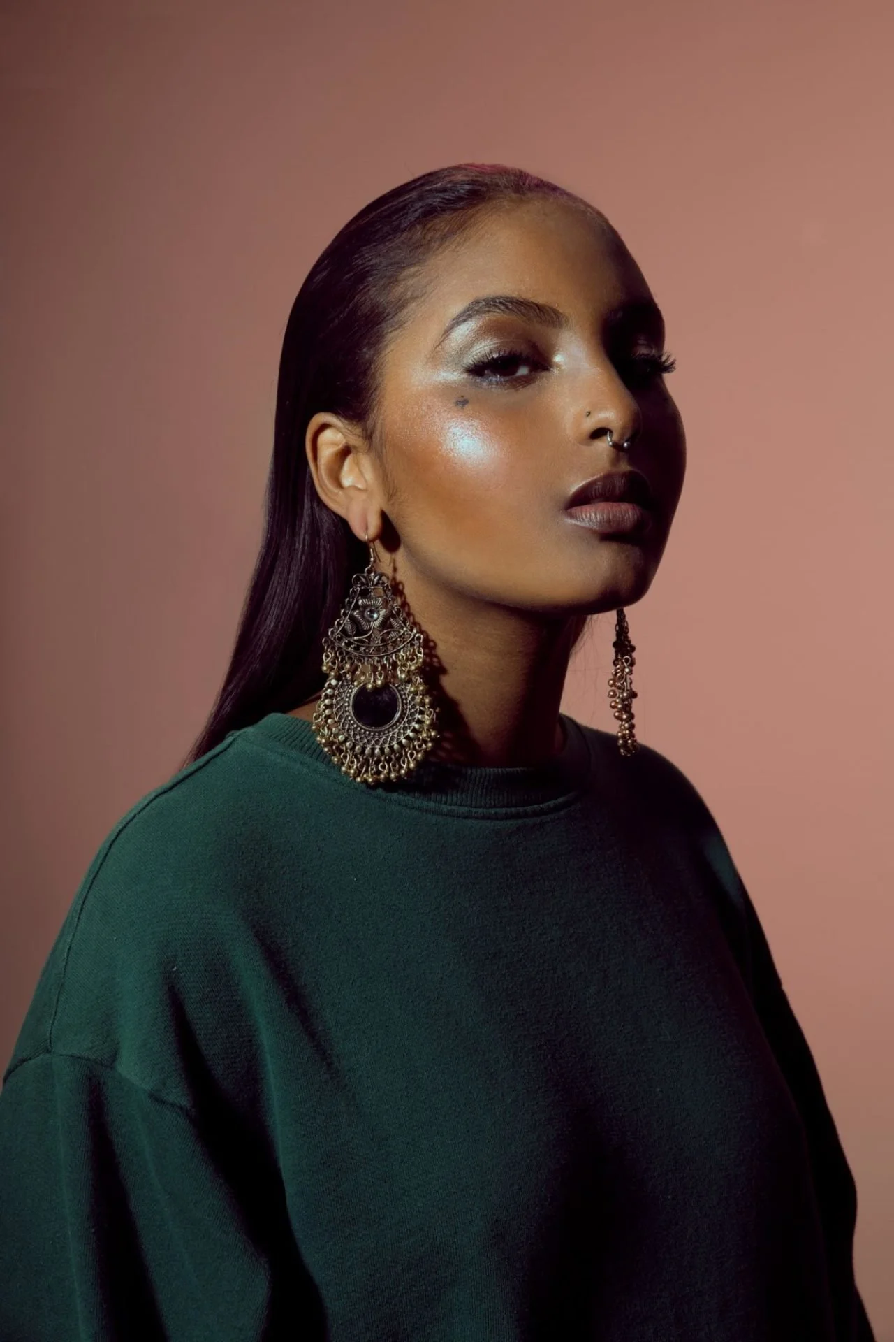 Un portrait d'une femme au teint foncé portant de grosses boucles d'oreilles ornementées et un sweat vert foncé, avec un fond rose clair.