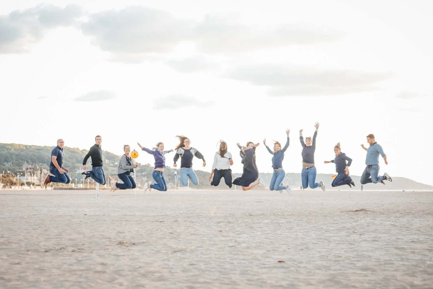 Groupe de personnes sautant en l'air sur une plage, avec un ciel nuageux derrière et un horizon urbain en arrière-plan.