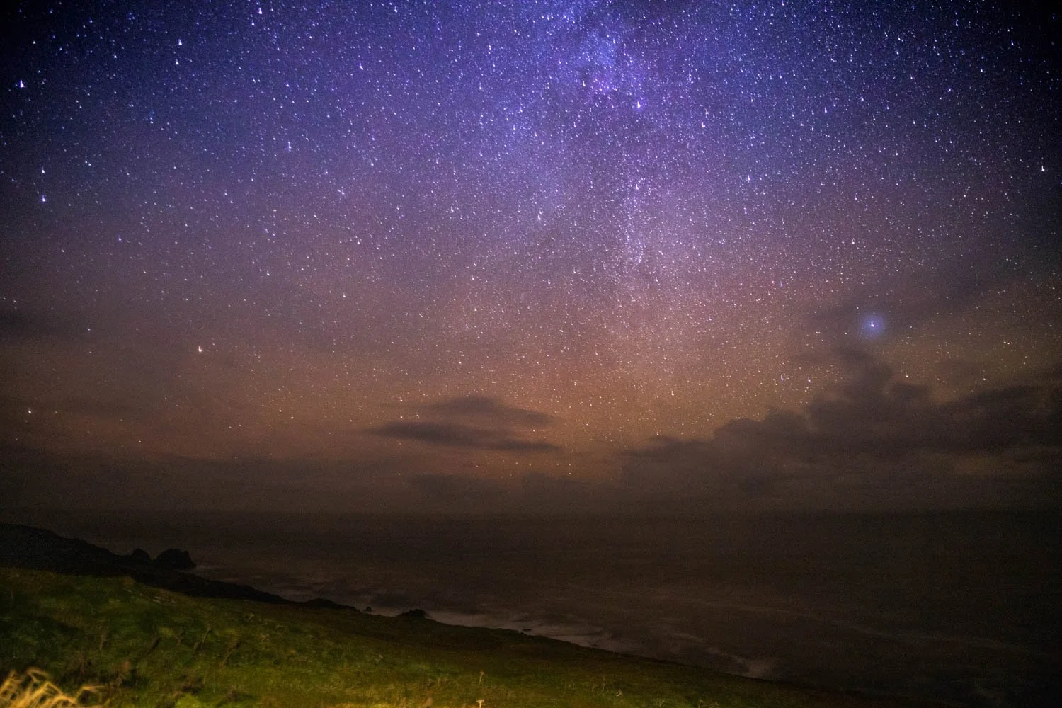 Ciel nocturne étoilé au-dessus de la côte avec des nuages bas et de la végétation verte en premier plan.