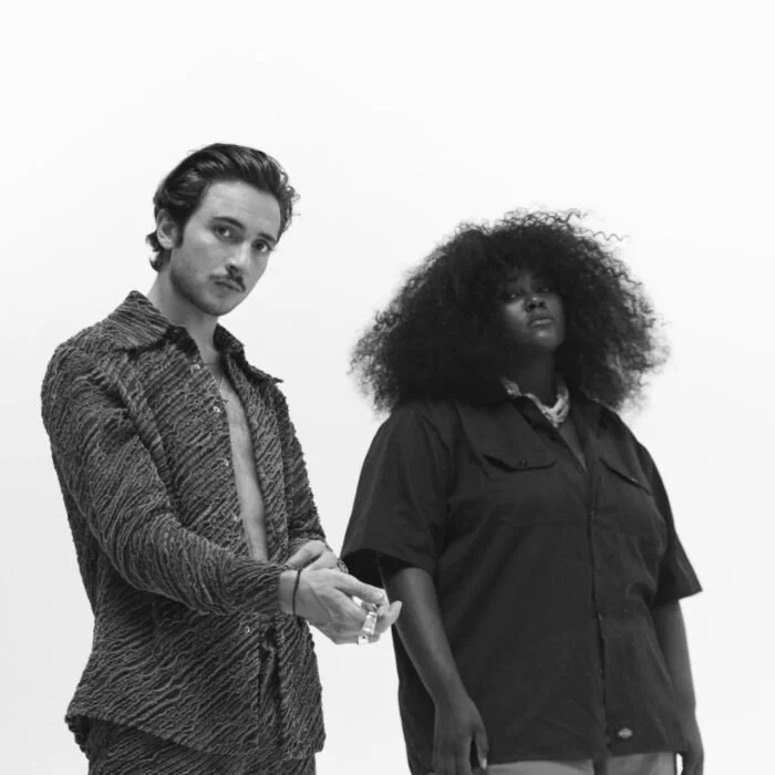 Deux personnes, un homme et une femme, posant côte à côte contre un fond blanc. La femme a de grands cheveux bouclés et porte une blouse foncée. L'homme a des cheveux courts et porte une veste texturée.