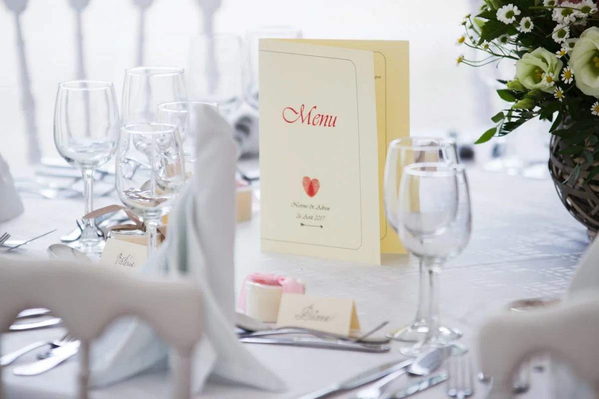 Une table de mariage élégante avec un menu, des verres, des assiettes, et un bouquet de fleurs blanches et vertes.