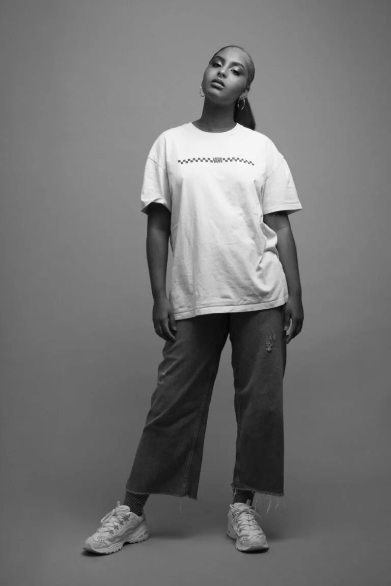 Jeune femme posant dans un studio avec un fond neutre, portant un t-shirt blanc avec un logo Vans, un jean large et des baskets, en noir et blanc.