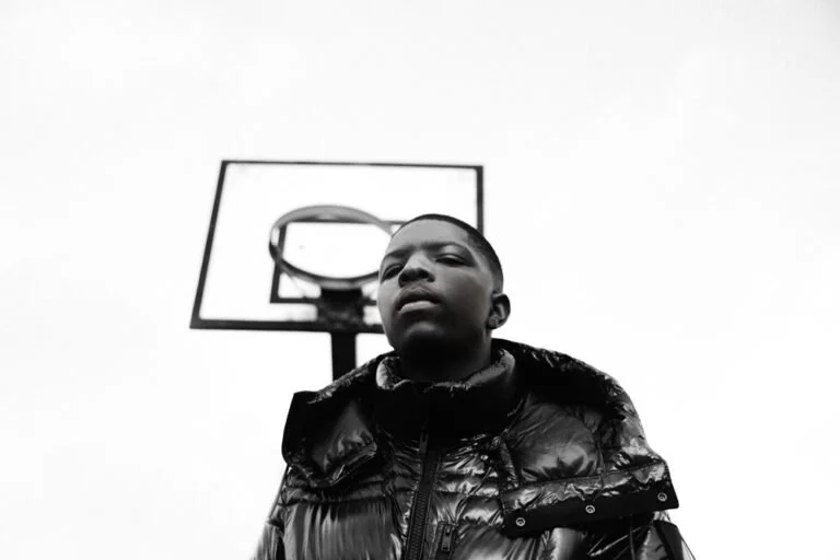 Jeune garçon en veste noire devant un panier de basket en plein air, photographié en noir et blanc.