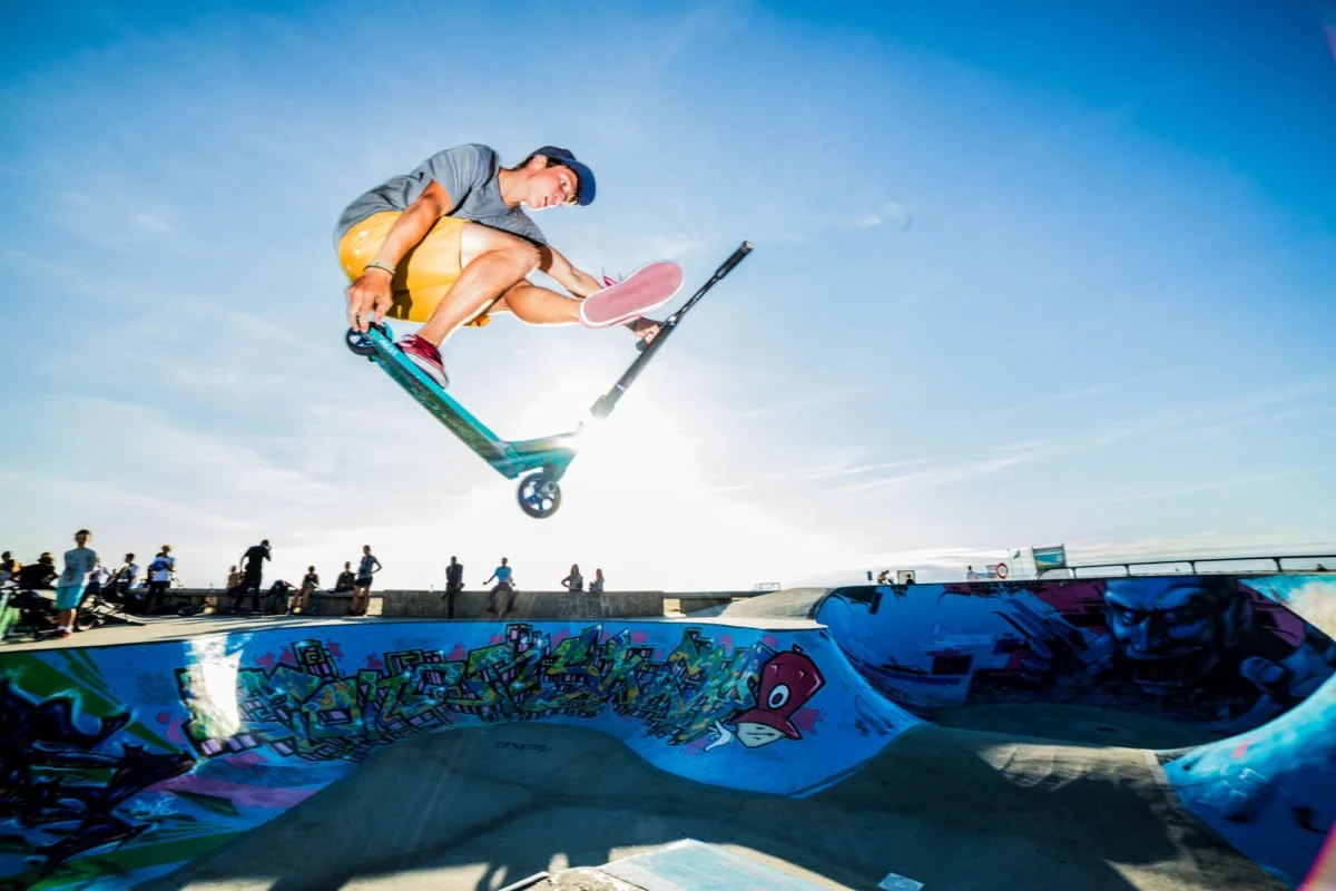Un skateur en l'air effectuant un saut dans un skatepark décoré de graffitis, avec un groupe de personnes en arrière-plan sous un ciel ensoleillé.